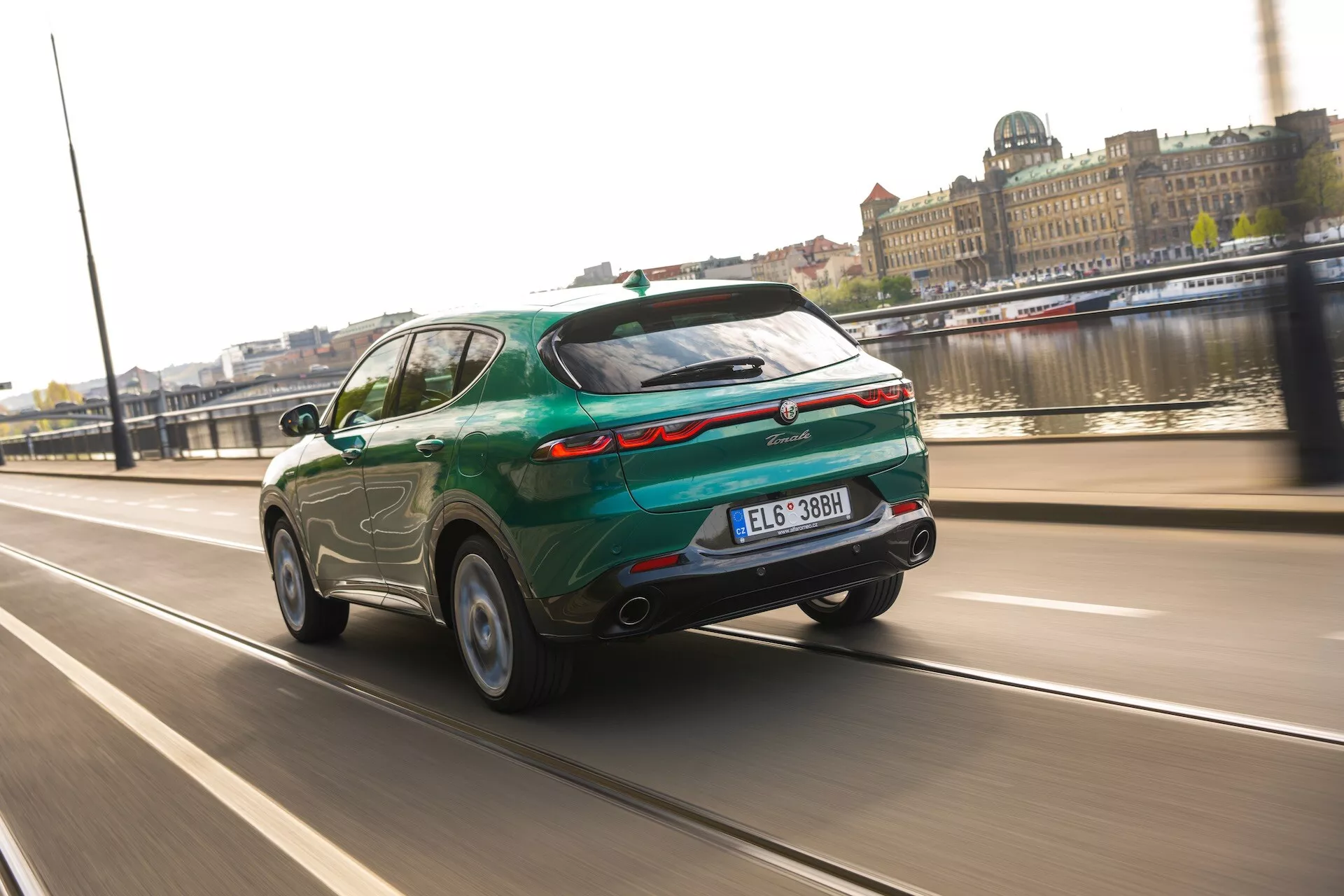 Moderen SUV Alfa Romeo Tonale 2022 v gibanju na mestni cesti z vodnim telesom in mestnimi zgradbami v ozadju, ki prikazuje poševeno streho, zadnji spojler, dvojni izpuh in zadnjo svetlobno palico po celi širini iz kota tri četrt nazaj.