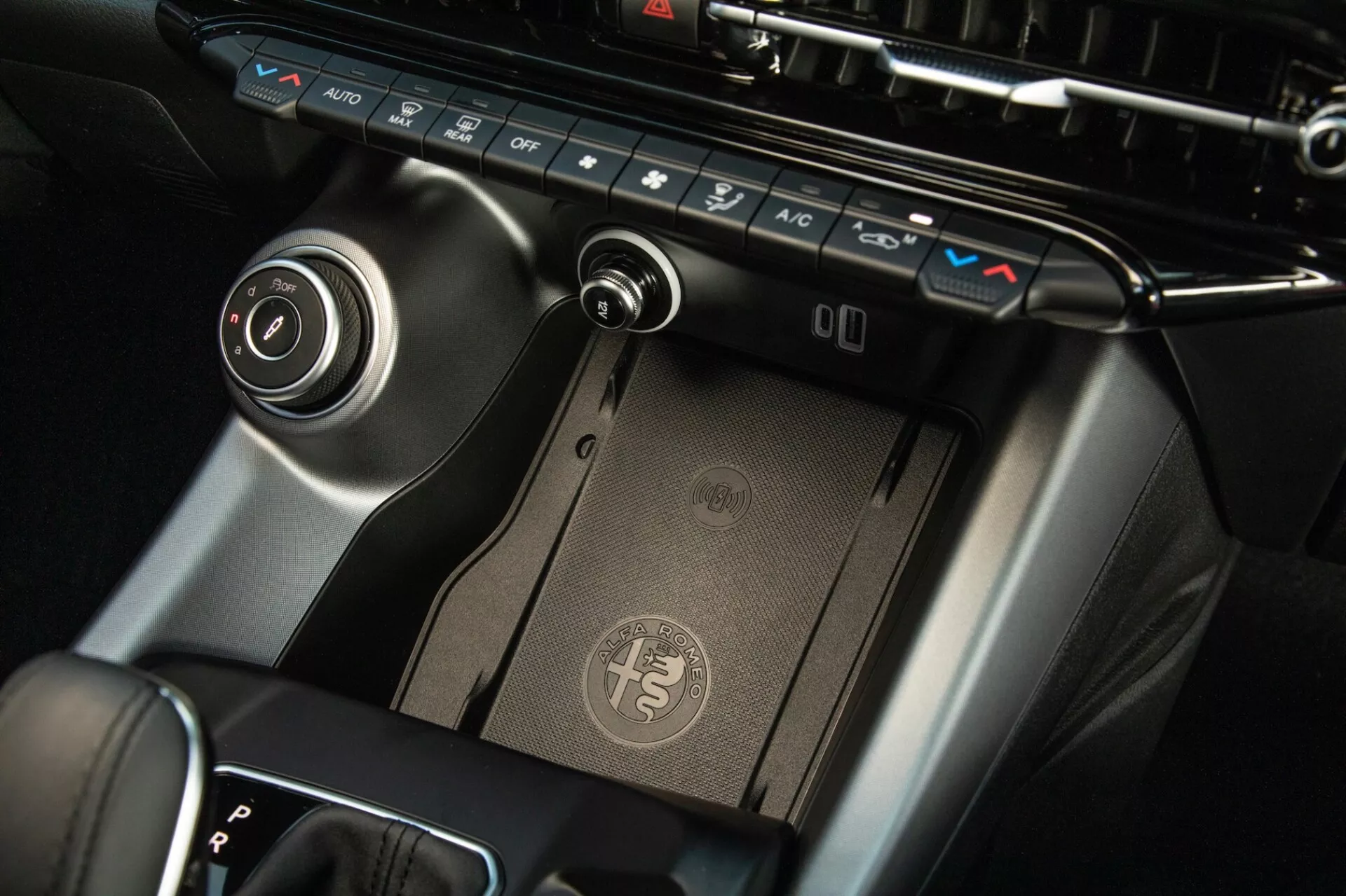 Podroben pogled na središčno konzolo Alfa Romeo Tonale 2022, ki prikazuje klimatski nadzorni plošči, brezžično polnilno ploščico, izbirnik prestav in krožno krmilno ploščo infotainment sistema z vrhunsko zaključno obdelavo in sodobno zasnovo.