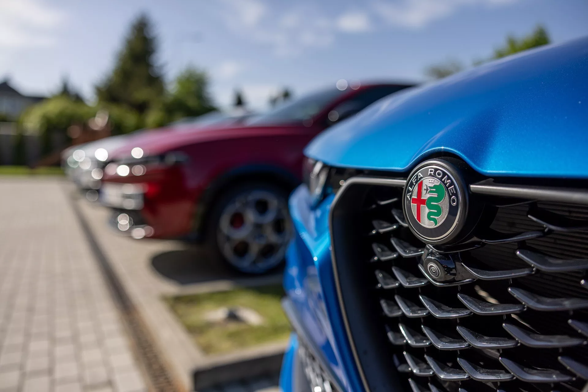 Podroben pogled na sprednjo rešetko modrega Alfa Romeo Tonale 2022 s poudarkom na emblemu, z navpičnimi lamelami in zamegljenim ozadjem drugih vozil