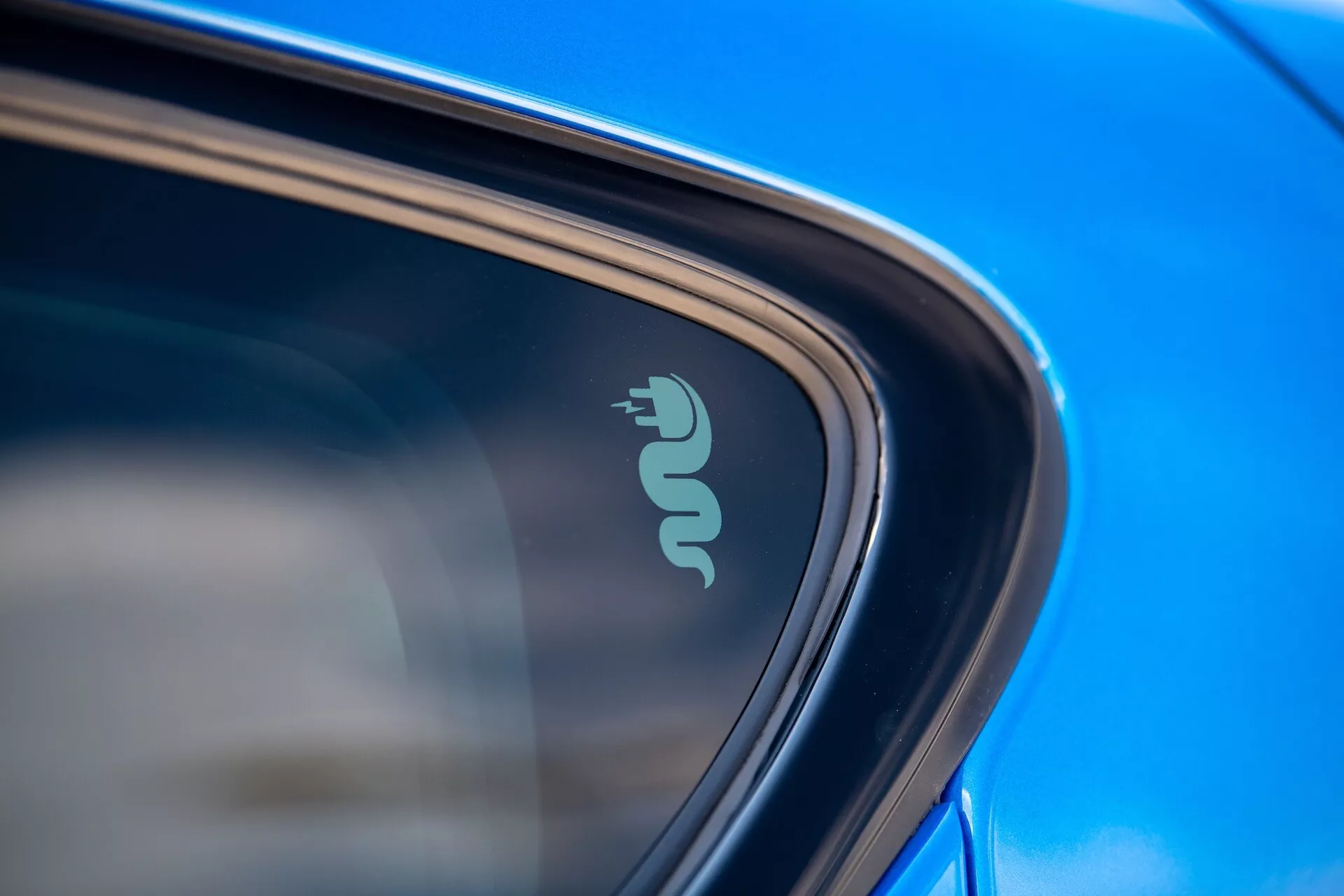 Podroben posnetek Alfa Romeo Tonale 2022, ki prikazuje tehnološko značko, ki nakazuje električno ali hibridno sposobnost blizu okna avtomobila, poudarjeno z odsevno modro barvo in elegantnim dizajnom v naravni dnevni svetlobi