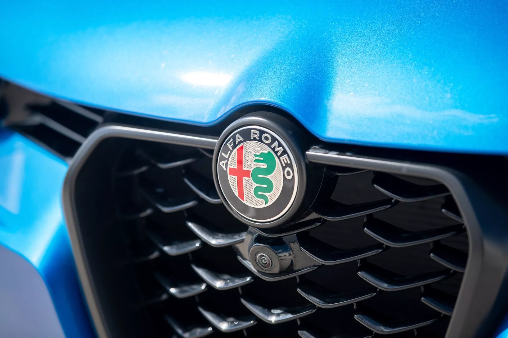 Podroben pogled na rešetko Alfa Romeo Tonale 2022, ki prikazuje ikonični znak z rdečim križem, zeleno kačo in modrim robom, osredotočen na izboljšano tehnologijo hlajenja in senzor za parkiranje