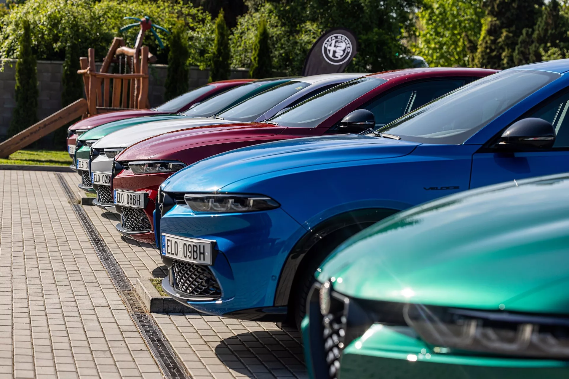 Lineup Alfa-Romeo Tonale iz leta 2022 v živahnih barvah prikazuje luksuzno oblikovanje z naprednimi LED žarometi, parkirani v diagonalni formaciji s poudarkom na sprednji levi strani iz nizkega do srednjega kota višine na tlakovani površini ob robu trave, morda pri trgovcu ali na dogodku ljubiteljev avtomobilov