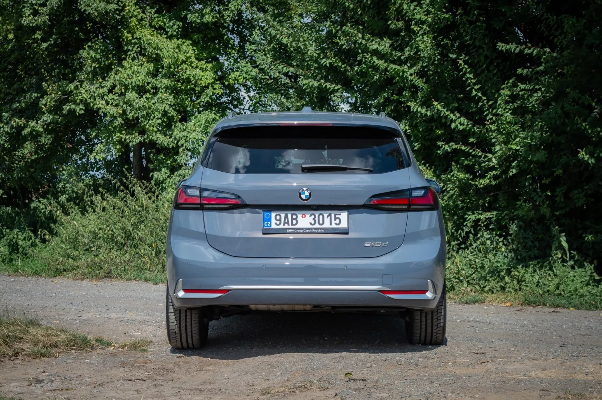 Zadnji pogled na BMW serije 2 Active Tourer 2022, poudarjajoč dvojne izpušne konice, senzorje za parkiranje zadaj in LED zadnje luči pod naravno osvetlitvijo z ozadjem podeželske zelenice.