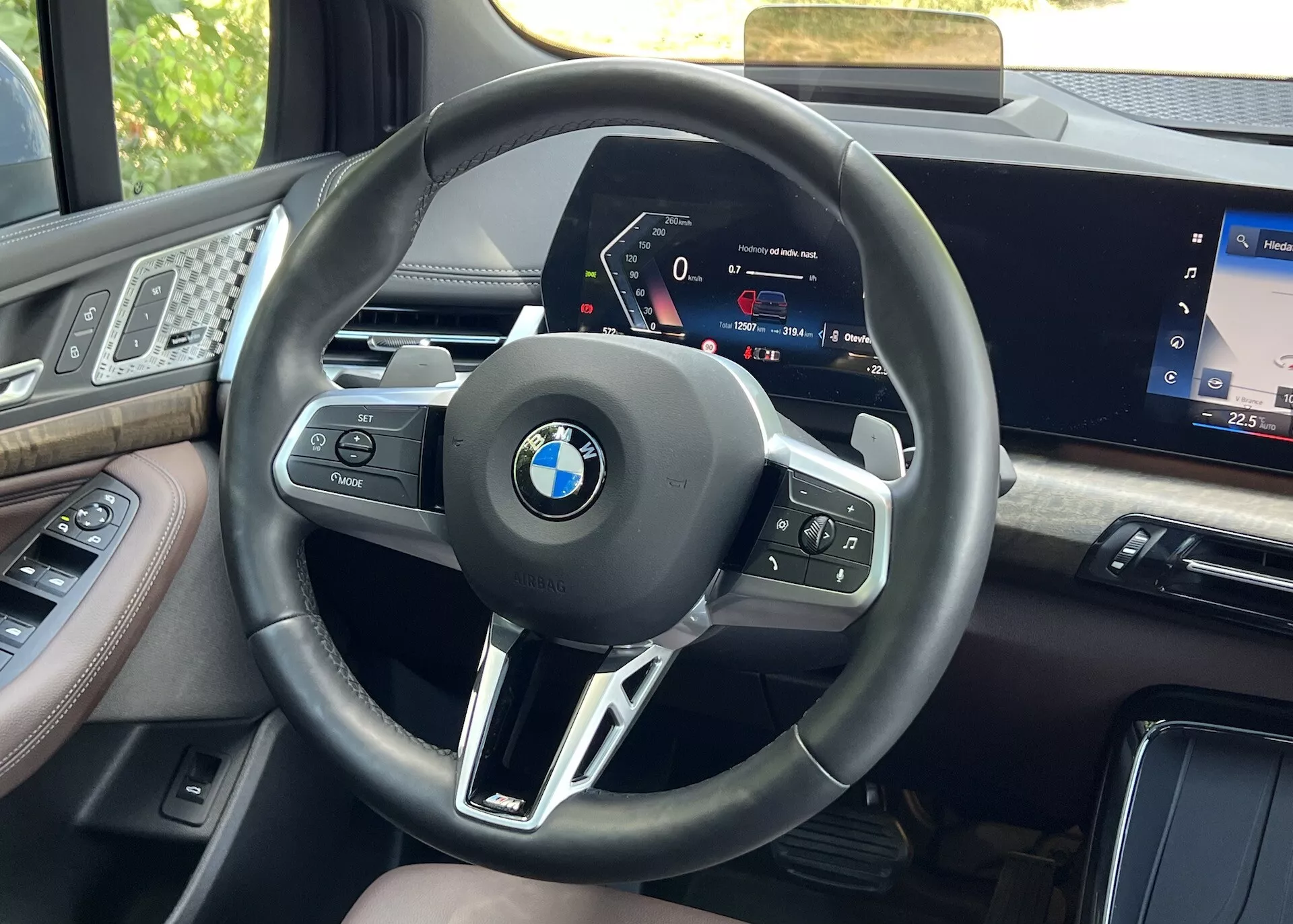 Pogled iz sedeža voznika notranjosti BMW serije 2 Active Gran Tourer letnik 2022, osredotočen na volan z večfunkcijskimi gumbi in visoko definicijsko digitalno instrumentno ploščo, obdan z visokokakovostnimi materiali in delom zaslona infozabavnega sistema