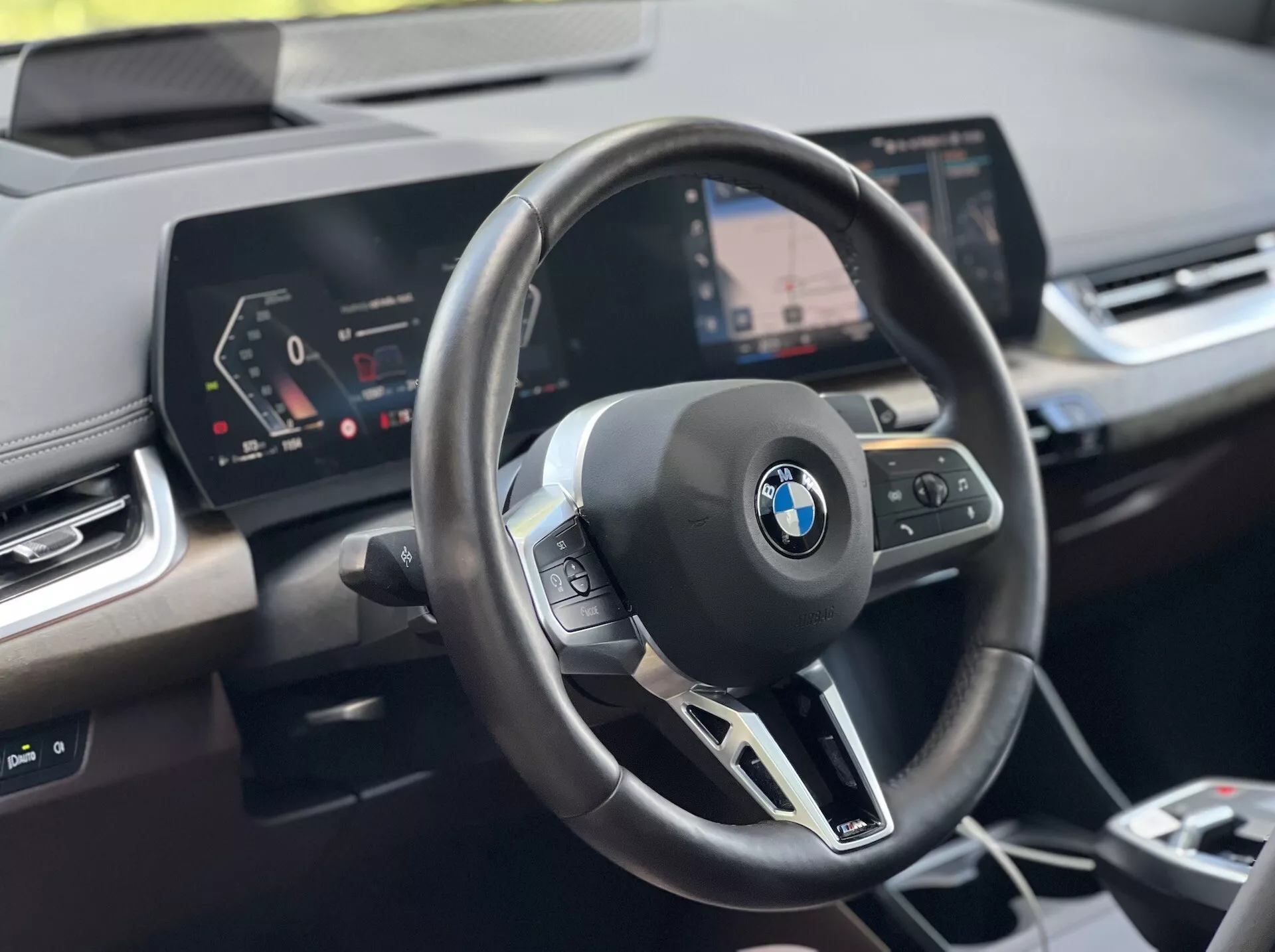 Notranji posnetek BMW serije 2 Active Tourer 2022: volan z emblemo BMW in večfunkcijskimi nadzornimi gumbi, digitalna instrumentna plošča in centralni infotainment zaslon z navigacijo, iz perspektive voznika