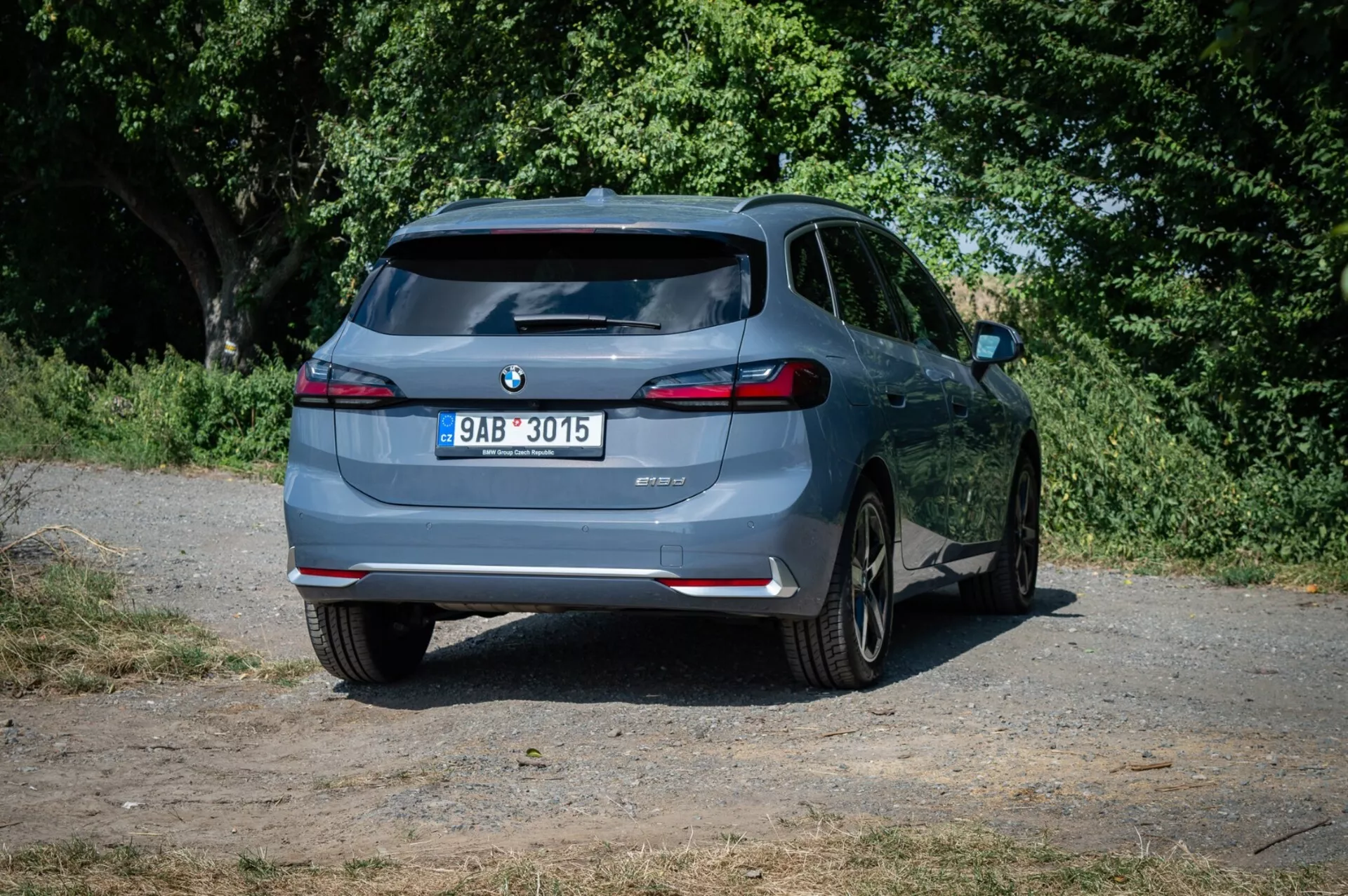 Hatchback BMW serije 2 Active Tourer 2022, pogledan z rahlo zamaknjenega zadnjega kota na podeželski cesti, poudarja njegov zadnji dizajn, LED zadnje luči, dvojni izpuh in tehnologijo pomoči pri parkiranju z naravnim ozadjem