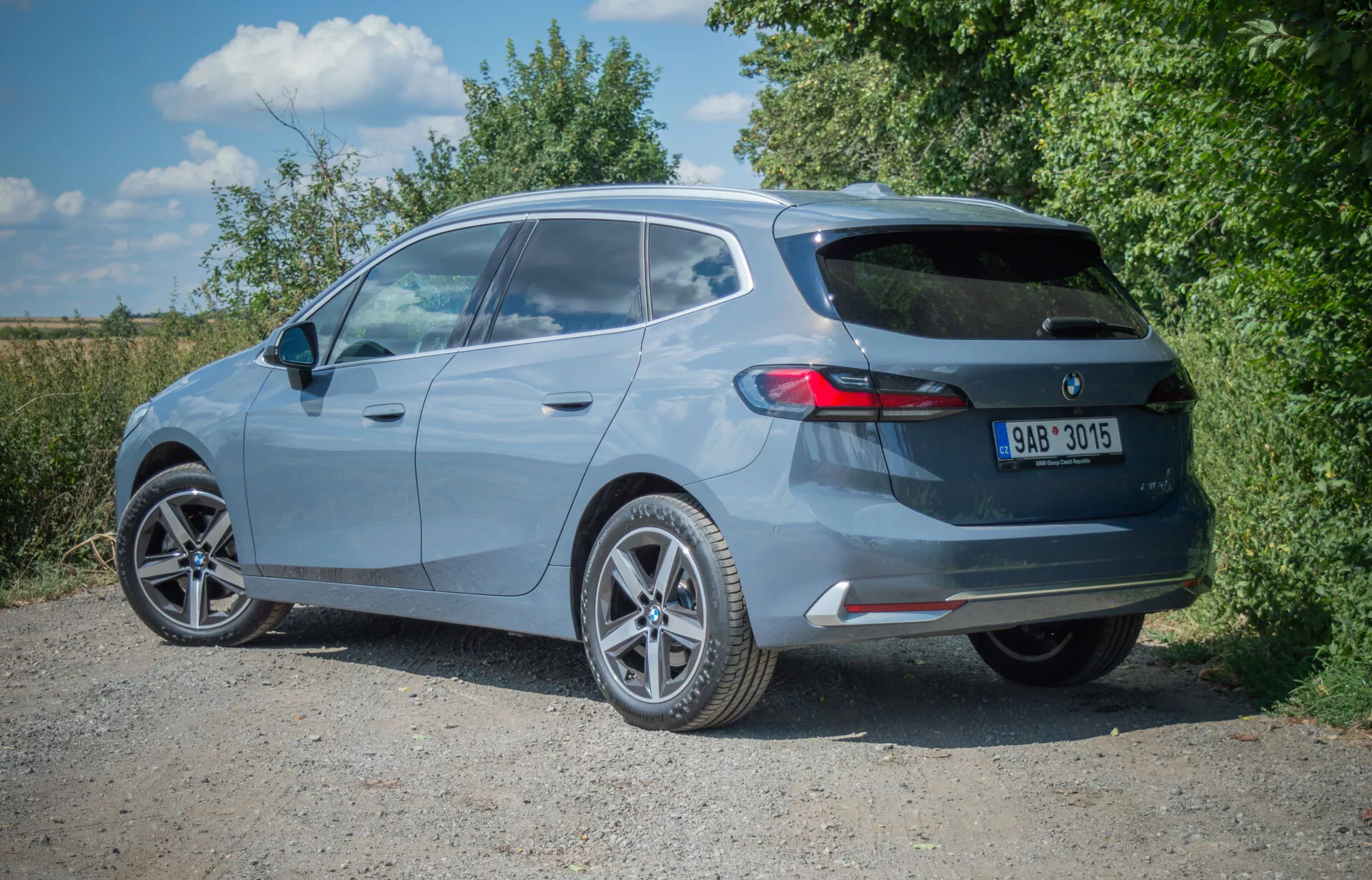 BMW serije 2 Active Tourer hatchback letnik 2022 v metalik barvi, zajet iz zadnje kotačne perspektive, prikazuje lita platišča, spojler na strehi, zadnji brisalec, tretjo zavorno luč, horizontalne zadnje luči, enojni izpuh in zadnji odbijač z integriranimi odsevniki, parkiran na neurejeni podlagi.