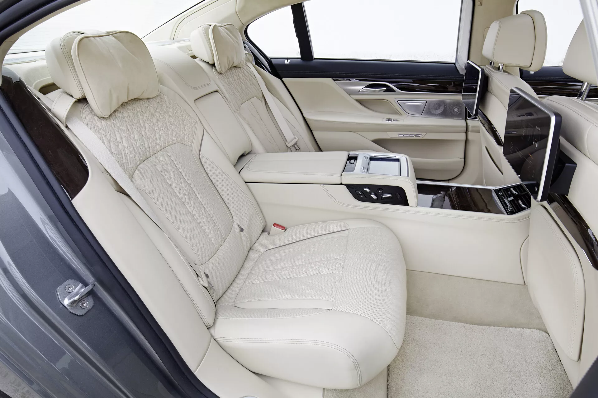 Notranjost BMW serije 7 (G11-G12) model 2015, ki prikazuje luksuzni zadnji del sedežev z usnjenim oblazinjenjem svetle kremne barve, oblikovani zadnji sedeži za dva potnika, ločeni s sredinsko konzolo. Konzola vsebuje tehnološke pripomočke, kot so nadzorna plošča, nadzor podnebja, ogrevanje ali hlajenje sedežev, možnosti medijev in držala za kozarce. Zasloni za zabavo zadaj so integrirani v zadnji del sprednjih sedežev s lesenimi obrobami na konzoli in vratih v svetlih svetlobnih pogojih.
