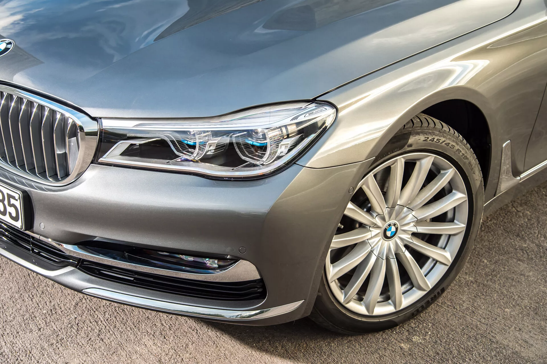 Bližnji poševen posnetek srebrnega luksuznega sedana BMW 7er-reihe 2015 (G11-G12), osredotočen na sprednji levi del vozila. Poudarjeni so sprednji odbijač, levi žaromet, pokrov motorja, blatnik in večkraka aluminijasta platišča. Kompleksni LED žarometi z značilno dnevno osvetlitvijo in majhnimi modrimi kvadratki, ki kažejo na napredne osvetlitvene sisteme. Delno vidna sprednja maska s kromiranimi navpičnimi letvami in kamera ali senzor v bližini nakazujeta tehnologijo za pomoč vozniku. Zračniki na sprednjem odbijaču in kovinska barva dodatno poudarjajo luksuzen in aerodinamičen dizajn vozila.