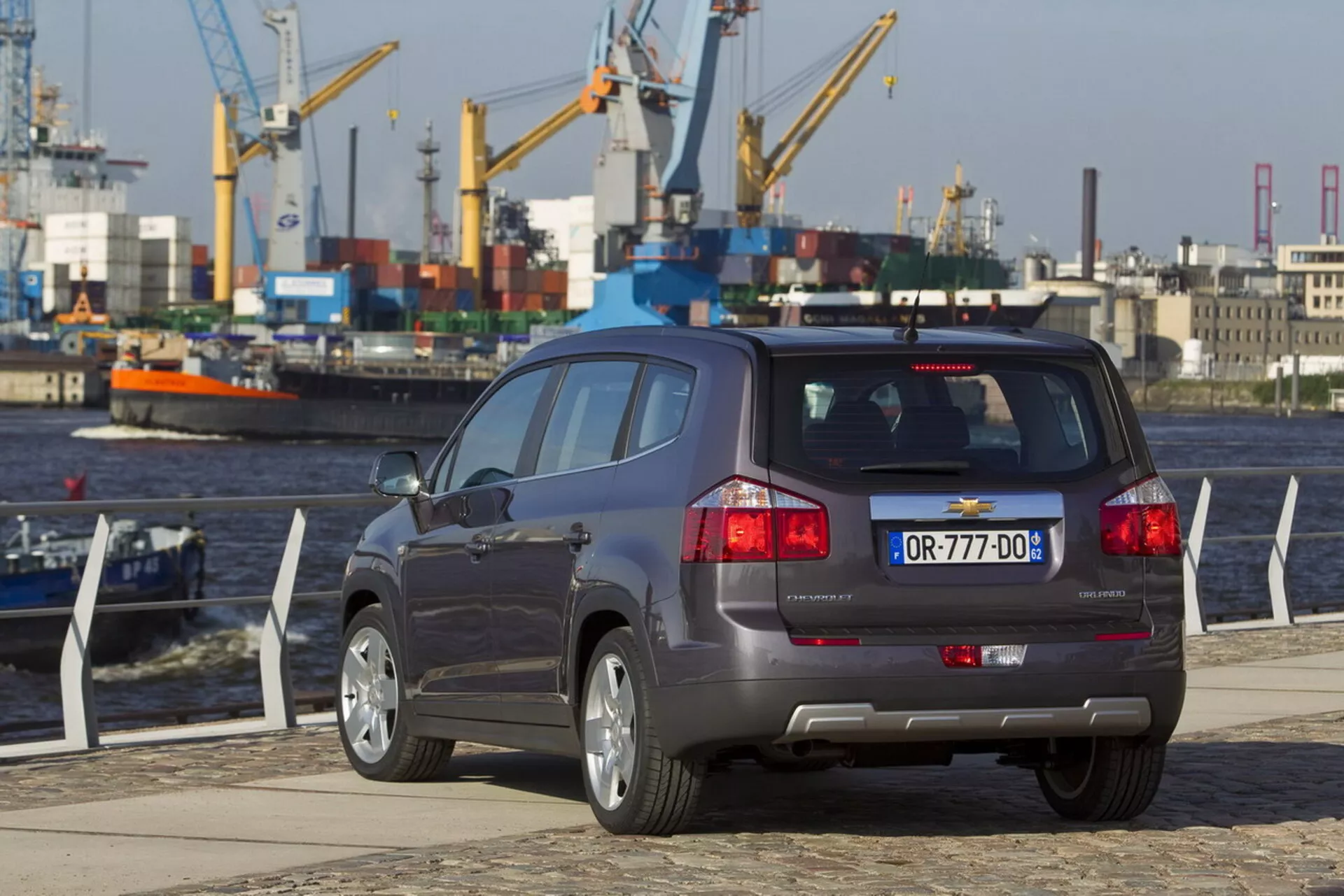 Zadnji tričetrtinski pogled na Chevrolet Orlando 2011 na pristanišču ob vodi, ki poudarja njegovo kompaktno SUV karoserijo, petvratno konfiguracijo, dvojni izpušni sistem, senzorje za pomoč pri parkiranju na odbijaču in večkraka lita kolesa