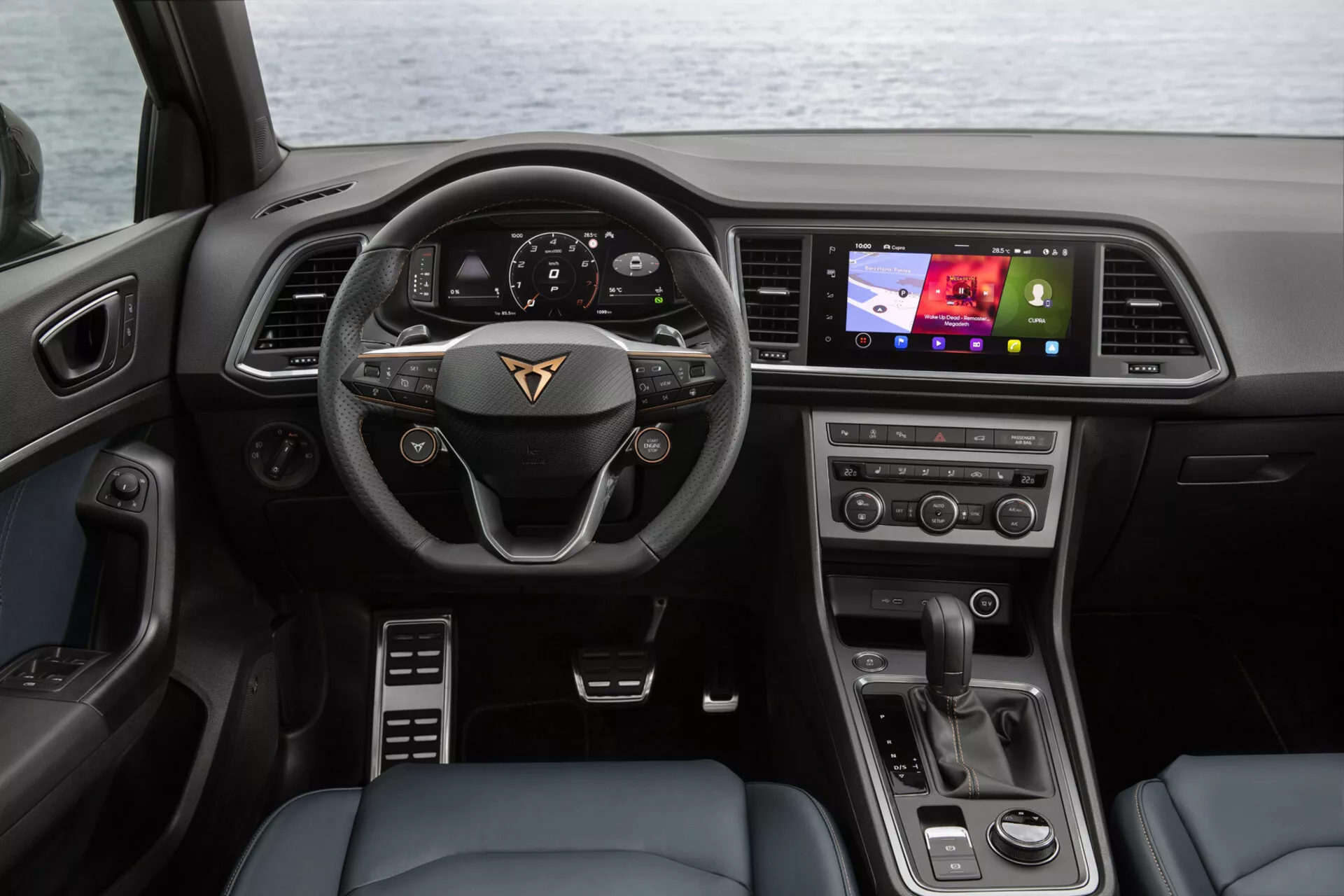Pogled na notranjost Cupra Ateca 2020 s sedeža zadaj s prikazom armaturne plošče, večfunkcijskega volana z emblemo proizvajalca, digitalnega instrument klasterja, na voznika usmerjenega zaslona na dotik za informacijsko-zabavni sistem, gumbov za nadzor klime in moderne izbirnika prestav