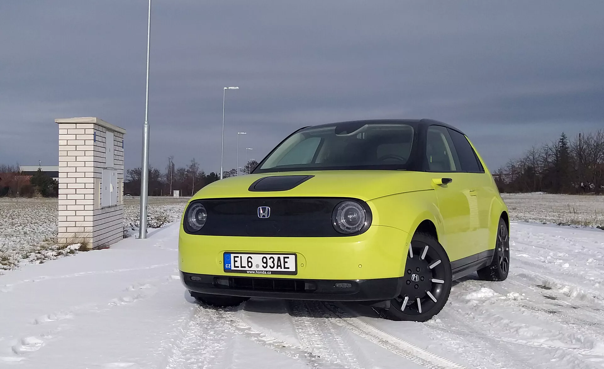 Honda e 2020 v svetlo rumeni barvi na snežno prekriti površini z oblačnim nebom, ki predstavlja LED žaromete, tehnologijo električnega vozila z naprednimi sistemi za pomoč vozniku, črna lita platišča in zimske pnevmatike.