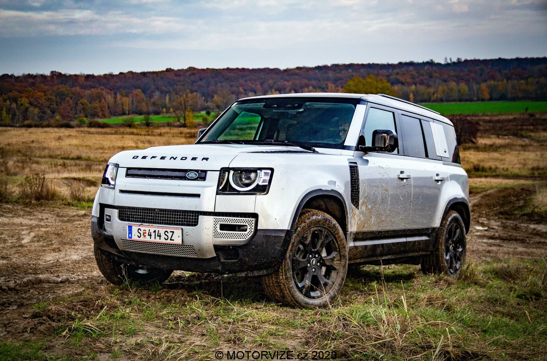 Slika terenskega vozila Land Rover Defender 2020 iz treh četrtin spredaj, postavljenega na odprtem polju z travo in drevesi v jesenskem ozadju. Robusten in škatlast dizajn vozila, prekrit z madeži blata, poudarja njegove terenske zmogljivosti. Značilne lastnosti vključujejo temno obarvana kolesa, napis 'DEFENDER' na pokrovu motorja, minimalistična sprednja rešetka z emblemom Land Rover, sodobne LED žaromete in robusten sprednji odbijač z zaščitnimi ploščami. Stopničasta strešna zasnova z alpskimi okni in velika zadnja potniška okna dodajajo funkcionalno estetiko avtomobila.