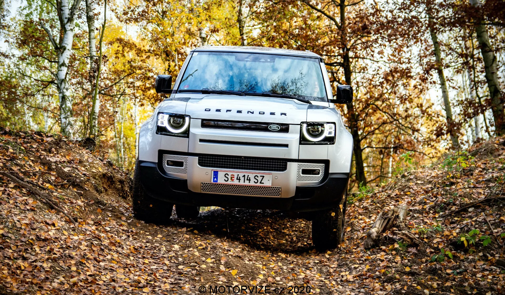 Čelni pogled na sodobni Land Rover Defender v naravnem jesenskem gozdu na neurejeni zemljani poti. Slika prikazuje mrežo, žaromete, pokrov motorja, kolesa in pnevmatike vozila, kar poudarja oddaljenost od tal in terenske zmogljivosti vozila. Ima sodobno LED osvetlitev, preprost dizajn mreže, senzorje na sprednjem odbijaču ali parkirno asistenco, jekleno zaščitno ploščo in avstrijsko registrsko tablico. Padajoče liste in drevesa z rumenim in rjavim listjem ustvarjajo sezonsko vzdušje. Vodni žig »MOTORYZE 2021« v spodnjem desnem kotu.