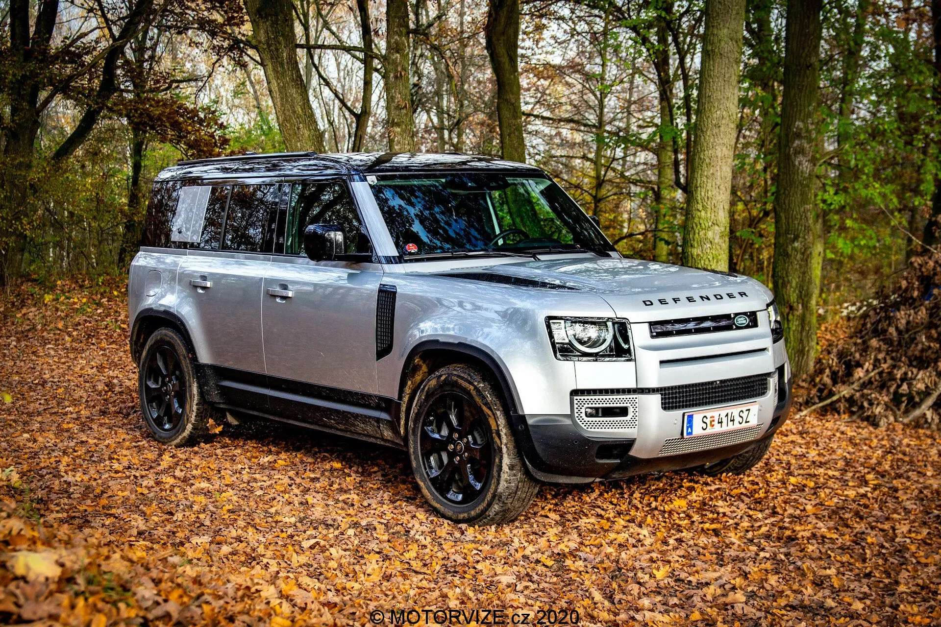 2020 Land Rover Defender iz tričetrtinskega sprednjega kota, ki prikazuje svojo robustno zasnovo in napredne funkcije v naravnem, jesenskem okolju z odpadlim listjem in drevesi. SUV ima srebrno karoserijo s črnimi podrobnostmi, LED žaromete, panoramsko streho in črna lita platišča. Slika zajema voznikovo stran in poudarja elegantno prednjo masko z logotipom DEFENDER, črna stranska ogledala z indikatorji in morebitne zaščitne plošče za vožnjo po terenu.