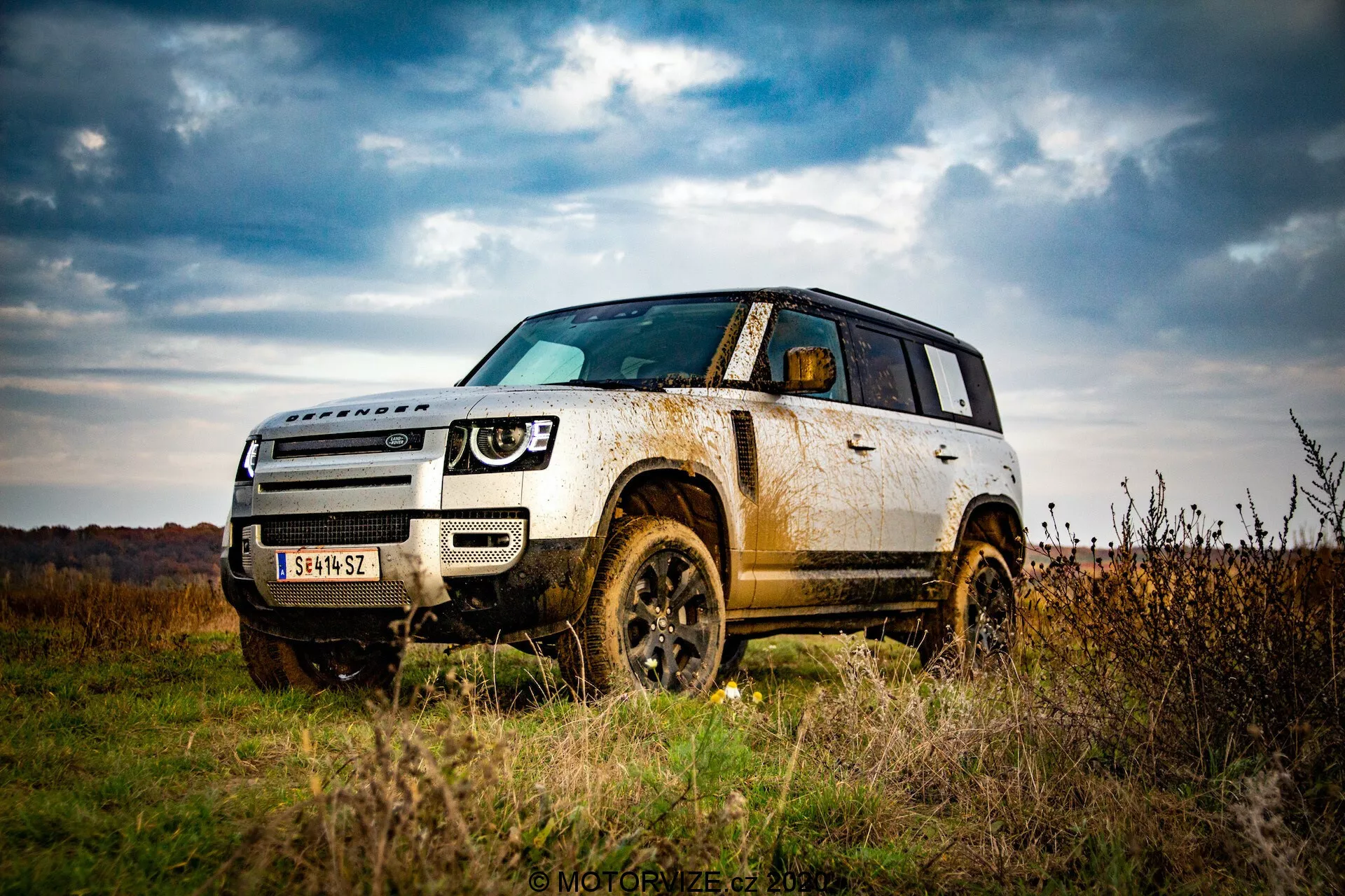 Podoba SUV Land Rover Defender 2020 na prostem, ki poudarja njegove terenske zmogljivosti z nizkega kota. Poudarja oddaljenost vozila od tal, njegovo robustnost z blatniki, pnevmatikami in zaščitnimi elementi na spodnjem delu karoserije. Pokaže pogled iz treh četrtin, prikazuje celoten sprednji del in stran do zadnjih vrat, poudarja oblikovalske elemente, kot so žarometi, mreža hladilnika in linije kontur na pokrovu motorja. Blato in umazanija na stranskih panelih in kolesih nakazujeta uporabo v terenu, kar kaže na vzdržljivost in vse terenske zmogljivosti. Oblačno nebo in jesenska pokrajina v ozadju povečujeta občutek pustolovščine.