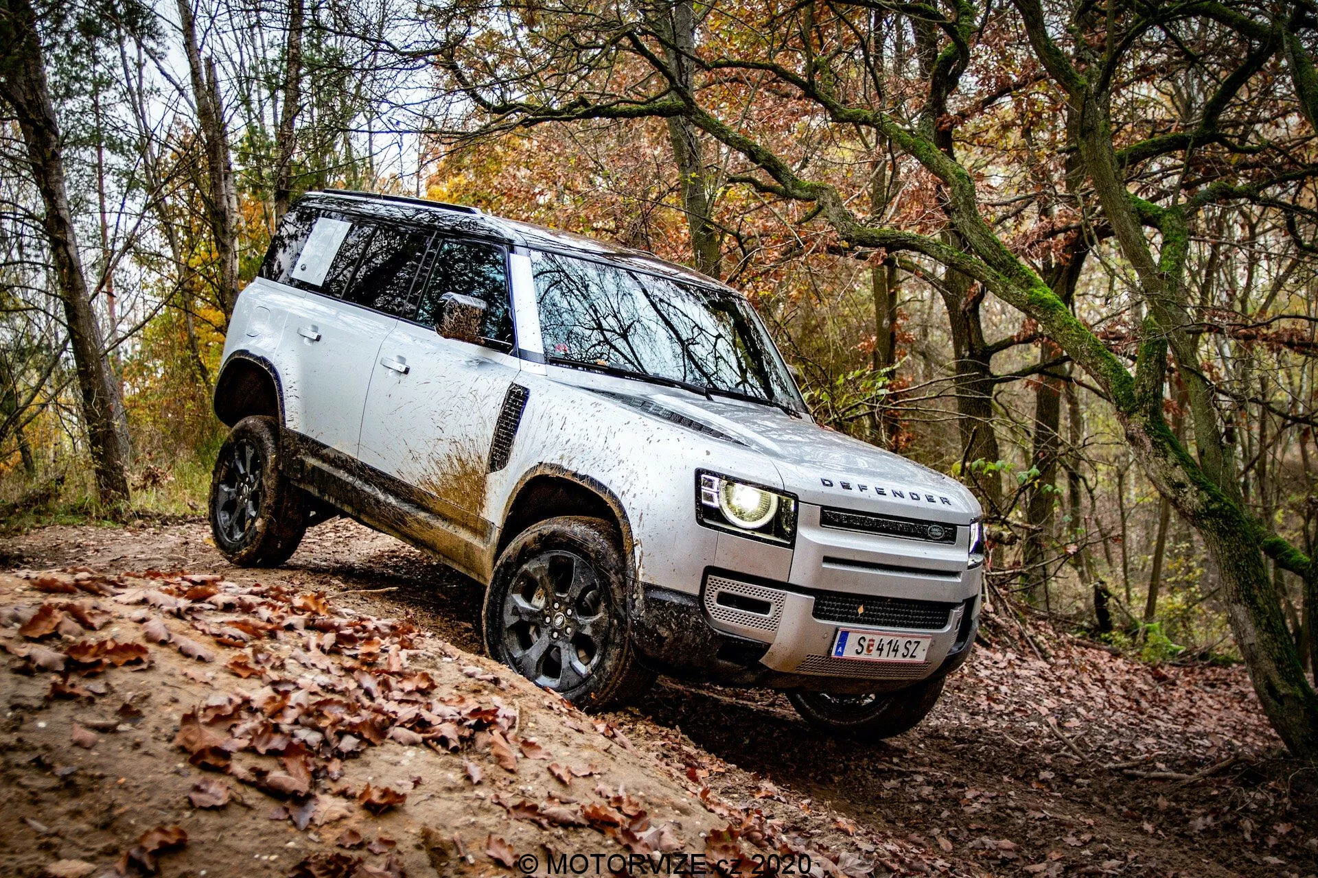 Stranski pogled na bel SUV Land Rover Defender 2020 v terenskem okolju; značilna zasnova z vidikom na sprednjo masko in odbijač, sodobne LED žaromete, črna lahka platišča s terenskimi pnevmatikami, nameščena stranska ogledala, škatlasta silhueta z blatom, poravnane kljuke vrat, ravna strešna linija in visoka oddaljenost od tal. Vozilo premaguje naklon, kar kaže na svoje terenske zmogljivosti.