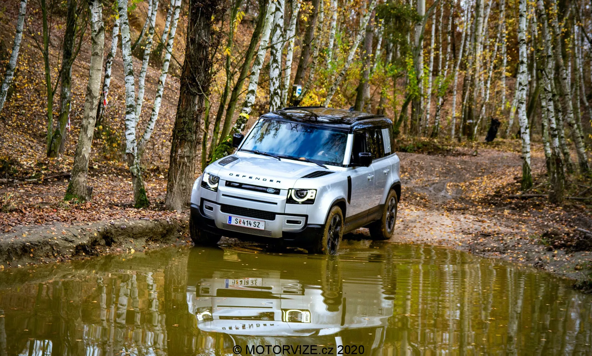 Slika Land Rover Defender 2020 v gozdnem okolju, zajeta iz sprednjega tritretjinskega kota. Vozilo prikazuje terenske zmogljivosti z vožnjo skozi blatno, z vodo napolnjeno pot. Oblikovanje vključuje sodobne žaromete, izrazito mrežo in zaščitno spodnjo ploščo. Strešni nosilci, pnevmatike z globokim profilom in odblesk vodne površine dodajajo estetski element. Okolje je obdano z brezami in padlim listjem, slika zajema bistvo jeseni.