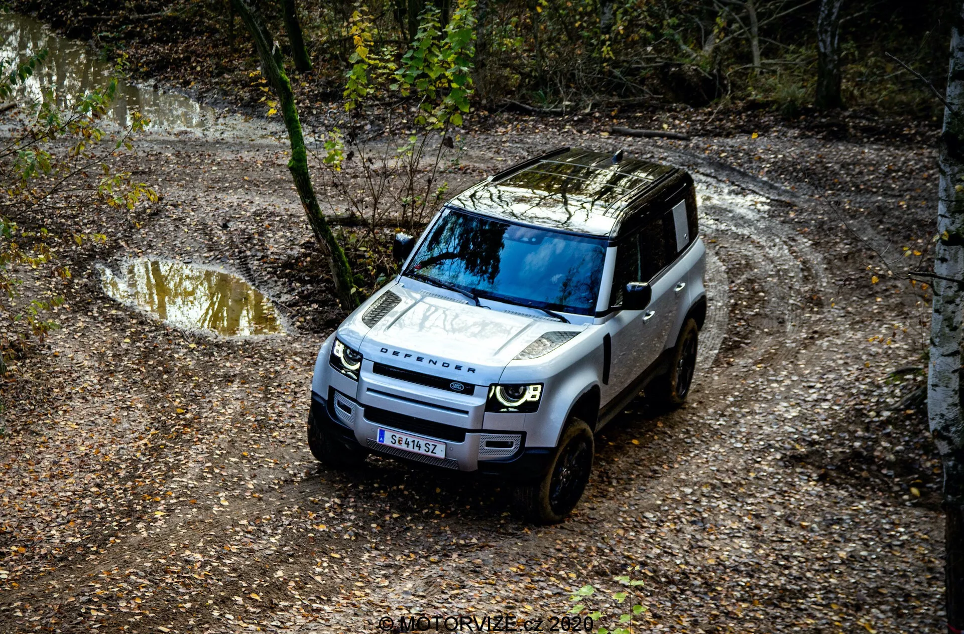 Pogled z višine na Land Rover Defender 2020, ki se vozi po terenski poti, z izrazitimi detajli, kot so prednja maska, pokrov motorja, vetrobransko steklo in del voznikove strani, obkrožen z naravnim okoljem z lužami vode in blata ter drevesi.
