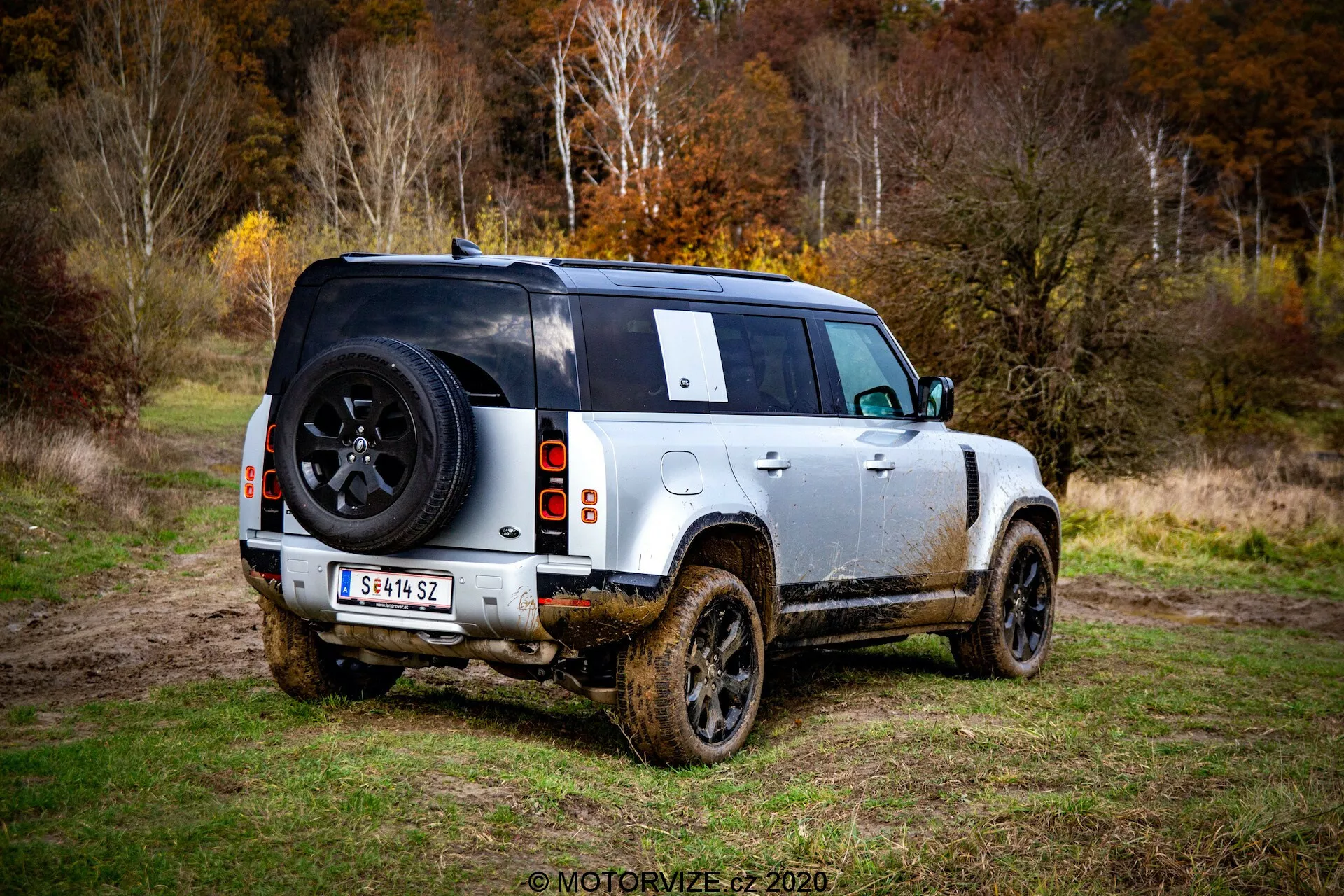 Zadnji trije četrtinski pogled na moderni Land Rover Defender 2020, štirivratni SUV, parkiran na travnatem območju z vidnim blatom ob straneh, kar kaže na nedavno uporabo v terenu. Fotografija, posneta s talne ravni, poudarja zadnje in bočne profile, osredotočena na zadnji dizajn, vključno z vidno zadnjo lučjo, zadnjim kolesom, prostorom za tovor in rezervnim kolesom, nameščenim na zadnjih vratih z logotipom znamke. Vozilo ima dvobarvno shemo z lahkim telesom in črno streho, aluminijasta platišča s trpežnimi terenskimi pnevmatikami, visoko oddaljenostjo od tal in delno zakrito zadnje okno z rezervnim kolesom ter zadnjim brisalcem. Blatne strani in spodnje plošče nakazujejo, da je bil SUV vožen po blatnem terenu. V ozadju je mogoče zaznati gozdnato območje z mehko svetlobo, kar kaže na oblačno nebo ali nizke svetlobne pogoje.