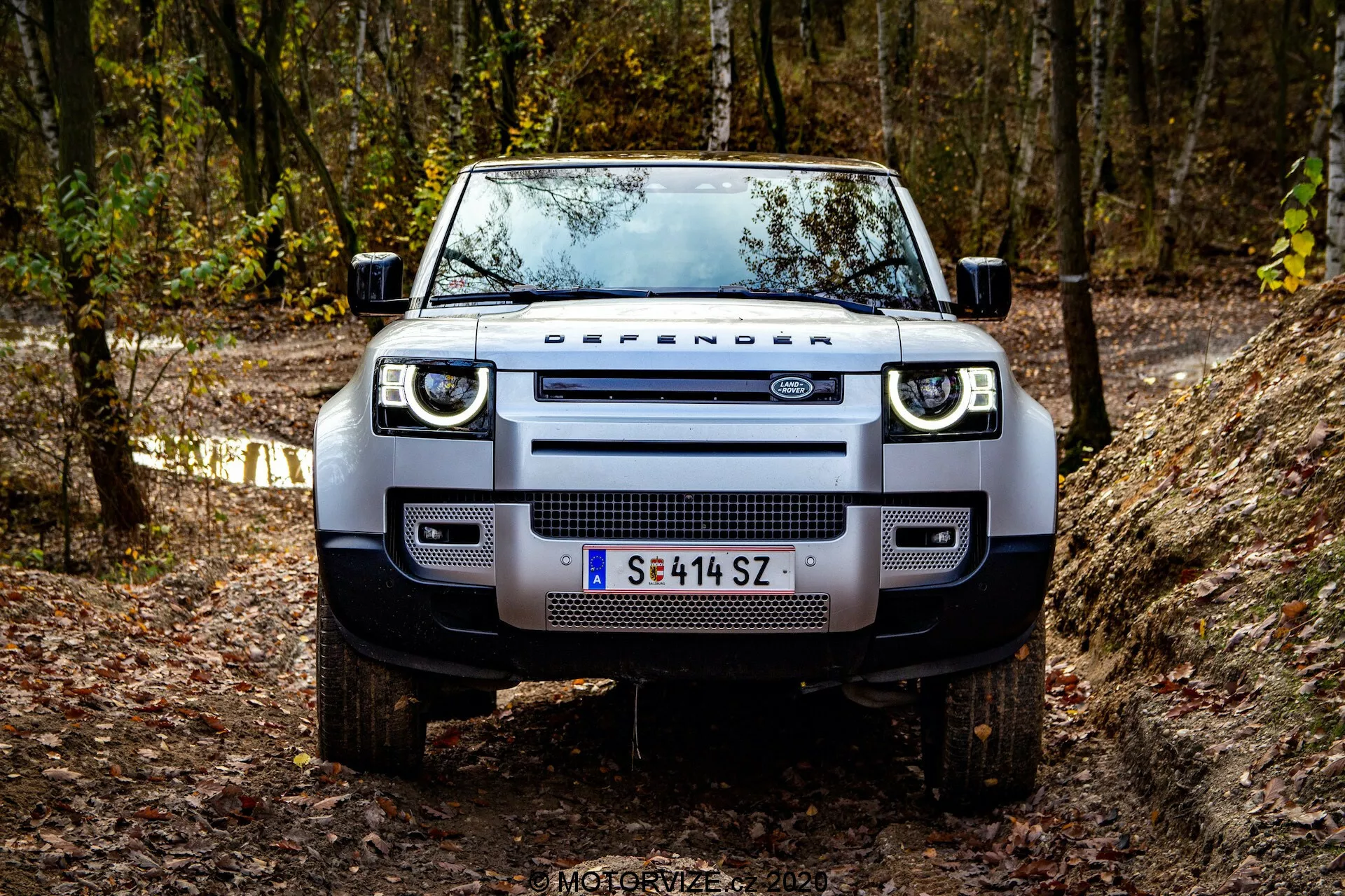 Frontalni pogled na Land Rover Defender 2020 na terenski poti, ki prikazuje njegovo trdno in dominantno prisotnost. Vidne podrobnosti vključujejo značilno mrežo s napisom 'DEFENDER', sodobne pravokotne žaromete z LED dnevnimi lučmi, širok sprednji odbijač z vgrajenimi meglenkami, robustne zaščitne plošče, ploščat pokrov motorja z izrazitimi robovi, masivne terenske gume na aluminijastih platiščih in funkcionalna stranska ogledala z integriranimi smerniki. Vozilo se nahaja v gozdnem območju z jesenskim listjem, kar poudarja njegov terenski in pustolovski duh.