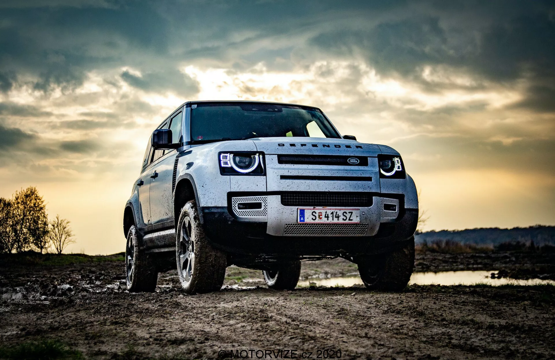 Slika prikazuje SUV Land Rover Defender 2020 parkirano na prostem, verjetno v terenskih pogojih. Posneto z nizkega kota poudarja robustno postavo in višino vozila. Sprednji trije četrti prikazujejo sprednjo masko, žaromete, pokrov motorja in blatnik ter kolo na voznikovi strani. Vidne so značilne LED dnevne luči, kar nakazuje na sodobne razsvetljavne funkcije. Drzna sprednja maska verjetno odraža identiteto proizvajalca, sprednja kamera ali senzor na maski pa nakazuje napredne sisteme za pomoč vozniku. Dramatično nebo v ozadju, verjetno z večerno ali mrakasto razsvetljavo, dodaja občutek avanture, medtem ko robustna kolesa in visoka razdalja od tal poudarjajo terensko sposobnost.