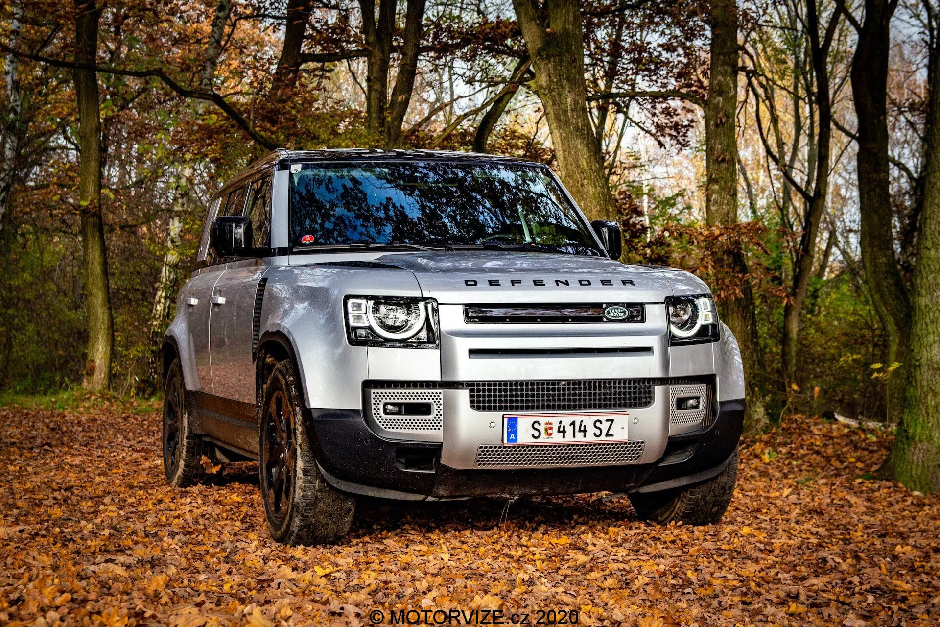 Pogled s sprednje strani pod kotom na sodoben Land Rover Defender 2020 v terenskem okolju z odpadlim listjem, posnet z nizkega kota. Slika poudarja sprednjo mrežo z napisom 'DEFENDER' na pokrovu motorja, LED žaromete s kvadratnimi dnevnimi lučmi in sodobne senzorje vgrajene v sprednji odbijač. Blatna kolesa in nastavljivo zračno vzmetenje nakazujejo terenske sposbnosti vozila. Vidna je stran voznika, sprednja vrata in del zadnjih vrat, prikazujoc robustno zasnovo vozila z delno zasukanimi kolesi.