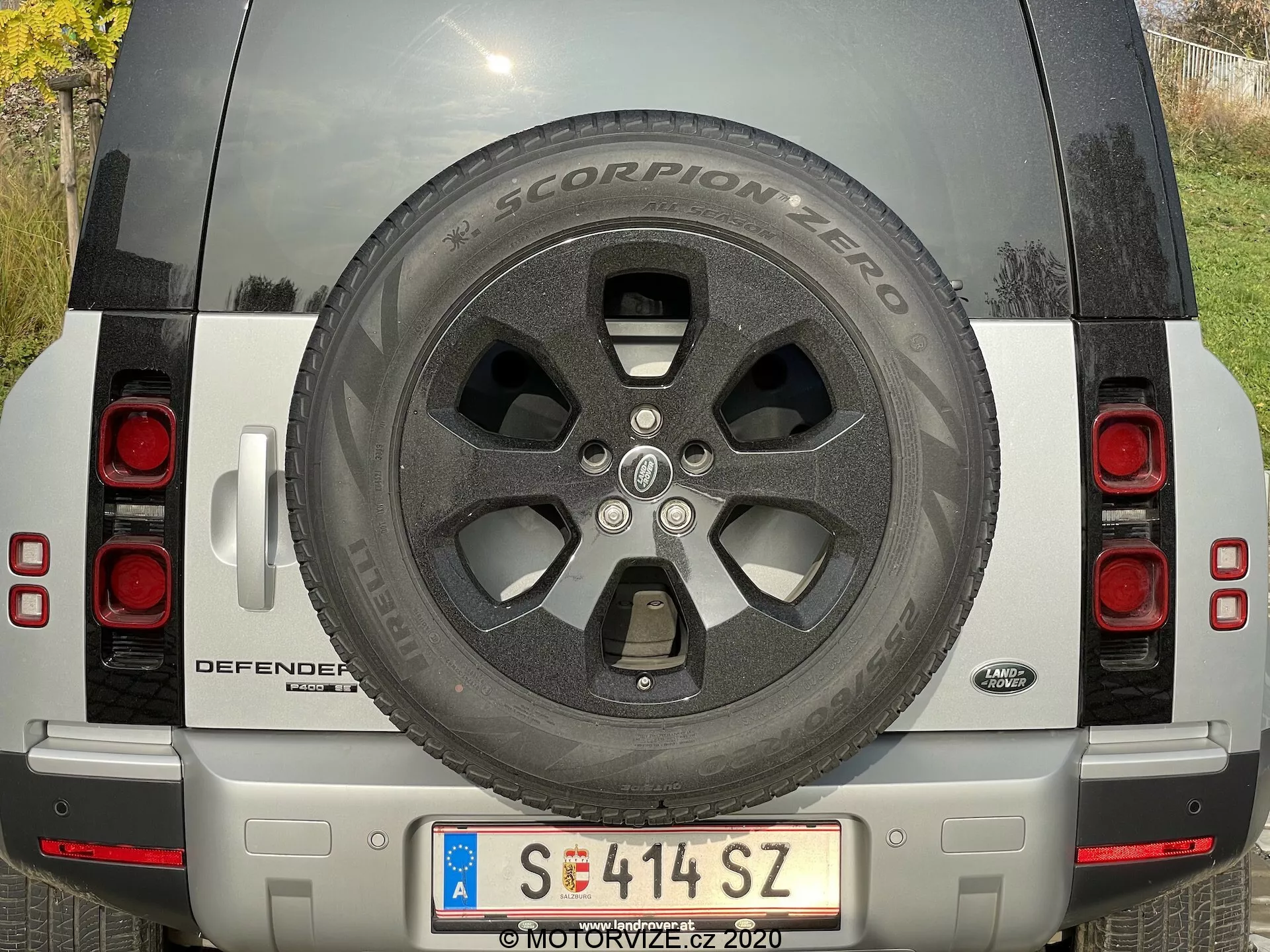Zadnji pogled na srebrni Land Rover Defender 2020 z vidnim rezervnim kolesom, zadnjimi lučmi, registrsko tablico, imenom modela DEFENDER in značko Land Rover, srebrnim odbijačem s senzorji in kamero ter odsevniki. SUV je parkiran v naravni dnevni svetlobi, kar prikazuje čisto in dobro vzdrževano stanje vozila.