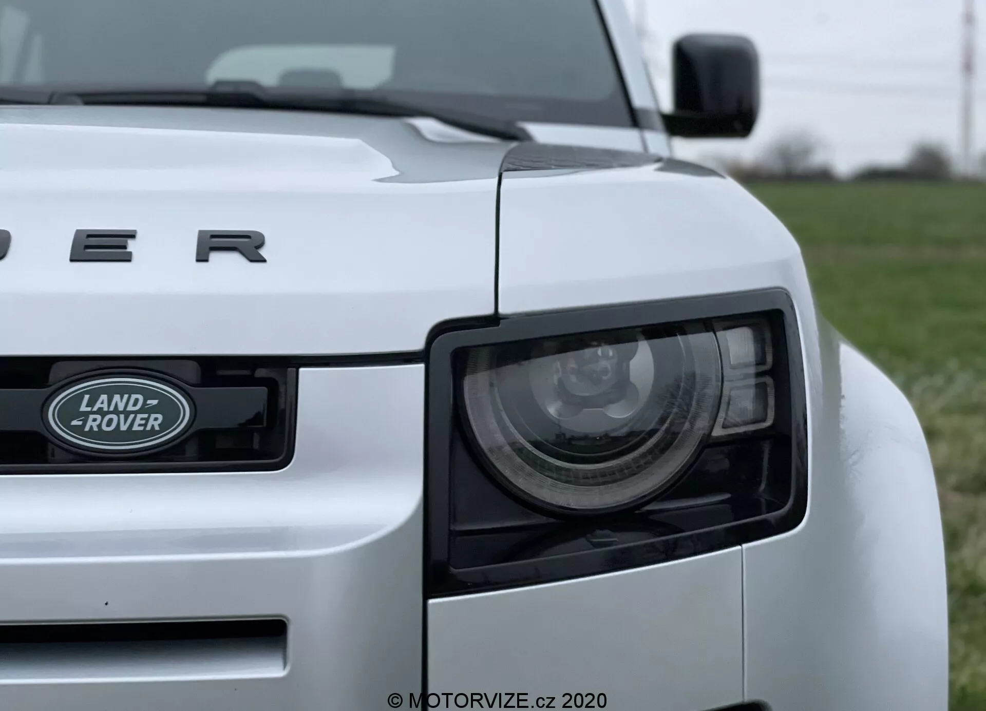 Povečan posnetek belega avtomobila Land Rover Defender 2020 iz nizkega kota, ki prikazuje levi sprednji kvadrant vozila. Vidni elementi vključujejo žaromet, rešetko z emblemom 'LAND ROVER' in pokrov motorja z napisom 'DEFENDER'. Žaromet vsebuje sodobne LED elemente, trdno zasnovo vozila pa poudarjajo vidne linije in krivulje. Avto je postavljen na travnato polje, z zabrisanim ozadjem, kar poudarja vozilo.