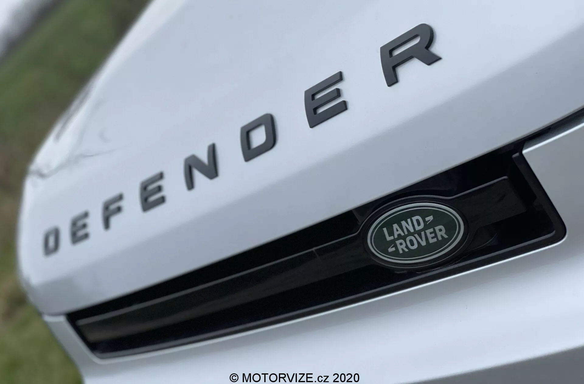 Bližnji pogled na sprednji del Land Rover Defenderja 2020, s poudarkom na oznaki 'DEFENDER' v dvignjenih srebrnih črkah na sprednjem robu pokrova motorja. Pod oznako je osrednje nameščena značka Land Rover v srebrno-zeleni barvi z belim robom na mreži. Slika je posneta iz nizkega, navzgor usmerjenega kota z majhno globino polja, kar poudarja elemente blagovne znamke, medtem ko je ozadje zamegljeno.
