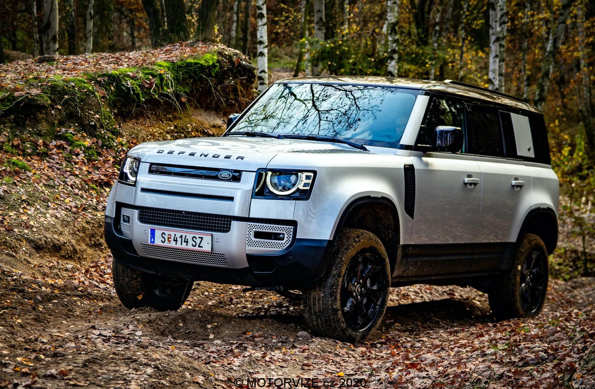 Nizek kotni posnetek Land Rover Defender 2020, posnet iz leve sprednje četrtine, poudarja robustne terenske lastnosti, kot so značilna sprednja maska Land Rover, LED žarometi z dnevnimi lučmi, zaščitna plošča na sprednjem odbijaču, izrazit DEFENDER branding na pokrovu motorja, kontrastna črna barva pokrova motorja na belem telesu vozila, povišani vstopi zraka za prečkanje globoke vode, veliki blatniki s črnimi terenskimi kolesi, smerniki na stranskih ogledalih in napredna vozniška asistenca v bližini sprednjega odbijača. Slika je nastavljena v terenskem okolju s padlim listjem, kar poudarja pustolovsko estetiko vozila. Vodni žig 'MOTORVIZA 2021'.