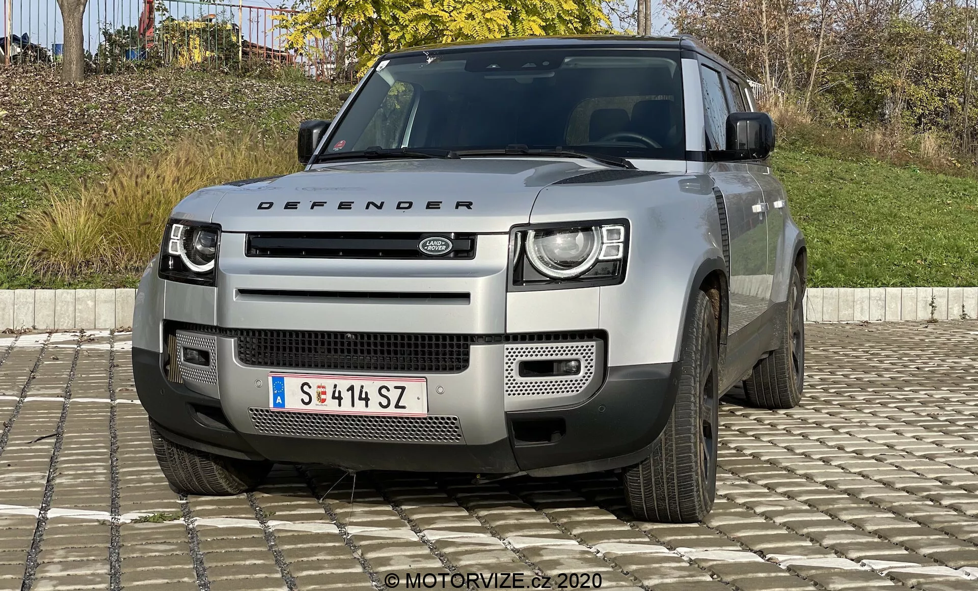 Prednji pogled na model Land Rover Defender 2020, ki poudarja njegove značilne dvo-stopenjske LED žaromete, izrazito masko z logotipom Land Rover, geometričen odbijač s srebrno zaščitno ploščo in druge robustne lastnosti, ki nakazujejo terenske zmogljivosti. Vidna sta tudi pokrov motorja z napisom 'Defender' ter zračna odprtina in delni pregled petkrakih koles. Vozilo je v svetlo nevtralni barvi, verjetno srebrni ali sivi.