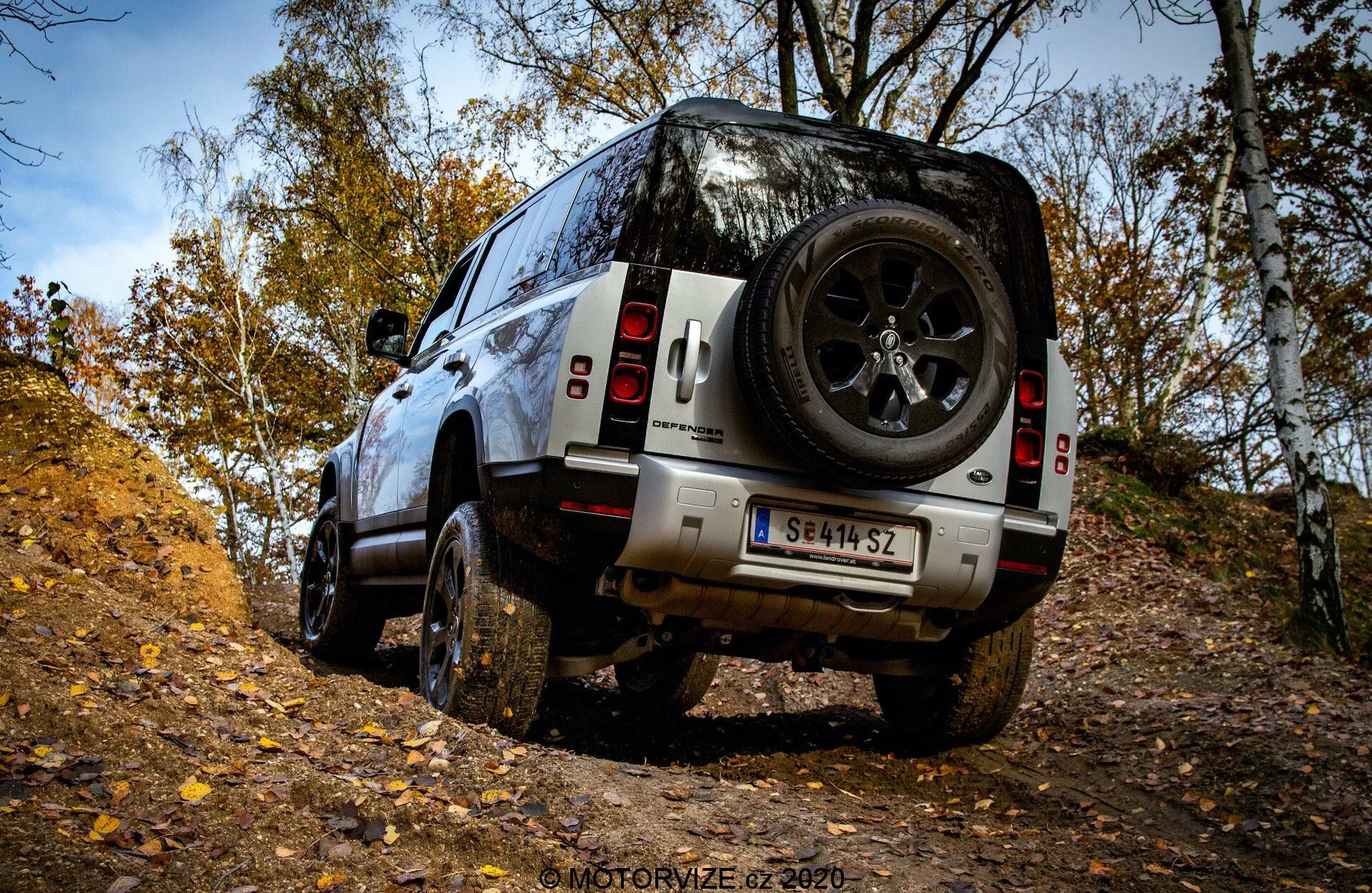 Pogled od zadaj v treh četrtinah na SUV Land Rover Defender 2020 na terenskih tleh, prikazuje robustne pnevmatike, veliko oddaljenost od tal in zadnjo rezervno kolo v jesenskem gozdnem okolju.