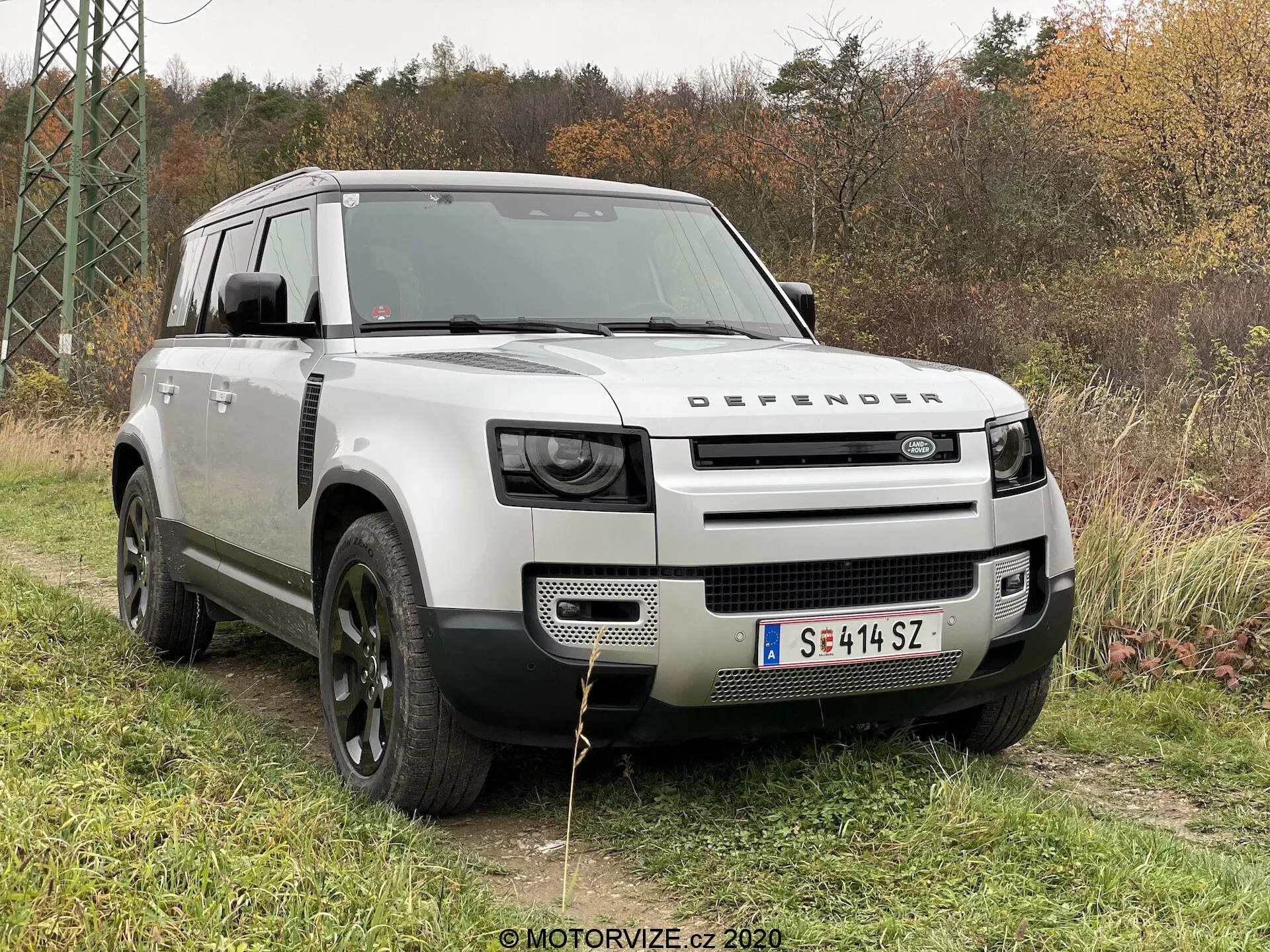 Ta slika prikazuje Land Rover Defender 2020 v zunanjem okolju na travi s listjem in drevesi v ozadju. Fotografiran s treh četrtinskim sprednjim kotom, fotografija poudarja izrazito mrežo z logotipom 'DEFENDER', sodobne LED žaromete, robusten pokrov motorja s školjkastim dizajnom, velika črna aluminijasta platišča s terenskimi pnevmatikami in srebrno karoserijo, ki kontrastira s črnimi blatniki in streho. Oblikovni elementi, kot so sprednja drsna plošča, bočna okna za dovod zraka 'alpine' in kvadratna zasnova, poudarjajo njegove terenske in uporabne zmogljivosti.