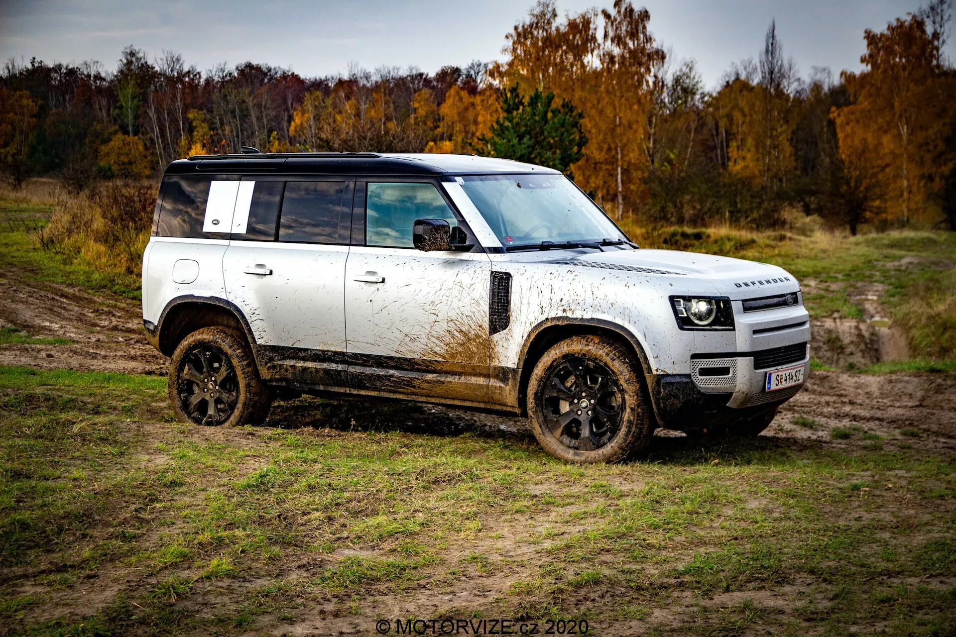 Stranski profil terenskega SUV-ja Land Rover Defender 2020, prikazan v tričetrtinskem kotu, z belo karoserijo s črno streho in kolesi, pokritimi z blatom. Vozilo je diagonalno postavljeno od spodnjega levega do zgornjega desnega kota proti blatnemu, naravnemu terenu pod oblačnim nebom z jesenskimi drevesi v ozadju.