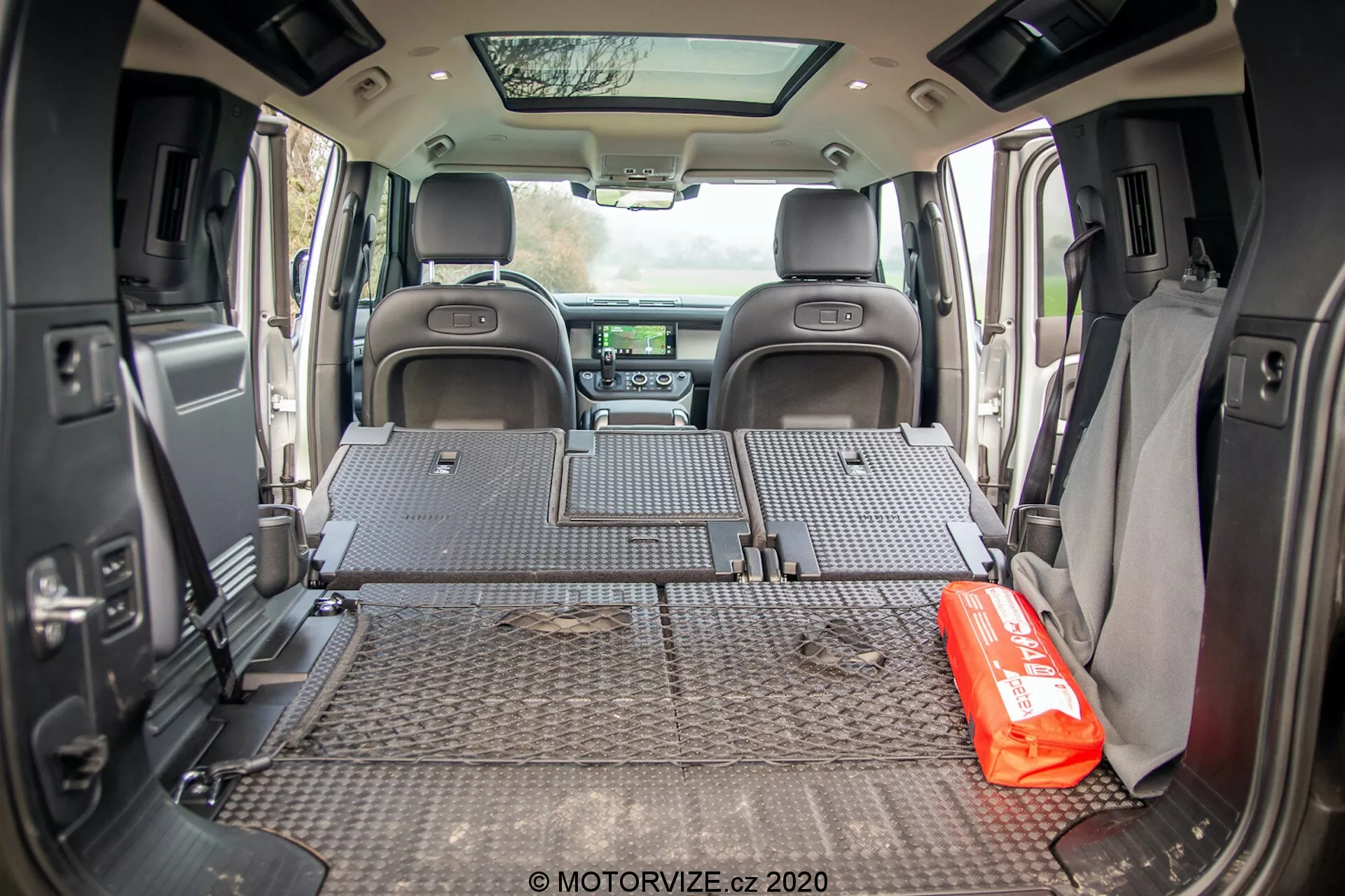 Notranji pogled prtljažnega prostora Land Rover Defender 2020, posnet iz zadnjega kota, prikazuje zaščitno preprogo z diamantnim vzorcem, ki pokriva celoten talni prtljažni prostor. Zadnji sedeži so zloženi plosko, kar razkriva prostoren prtljažni prostor. Stranske stene imajo različne pripomočke, kot so zvočniki in nosilci za kozarce, kar nakazuje na uravnotežen dizajn med funkcionalnostjo in udobjem. Oddaljena sprednja armaturna plošča je delno vidna in vsebuje informacijsko-razvedrilni sistem in nadzor podnebja. Na tleh prtljažnega prostora je izrazito postavljena rdeča kompleta za nujne primere, kar nakazuje na družinsko ali avanturistično uporabo vozila. Odprta zadnja vrata omogočajo, da naravna svetloba prodira v prtljažni prostor.
