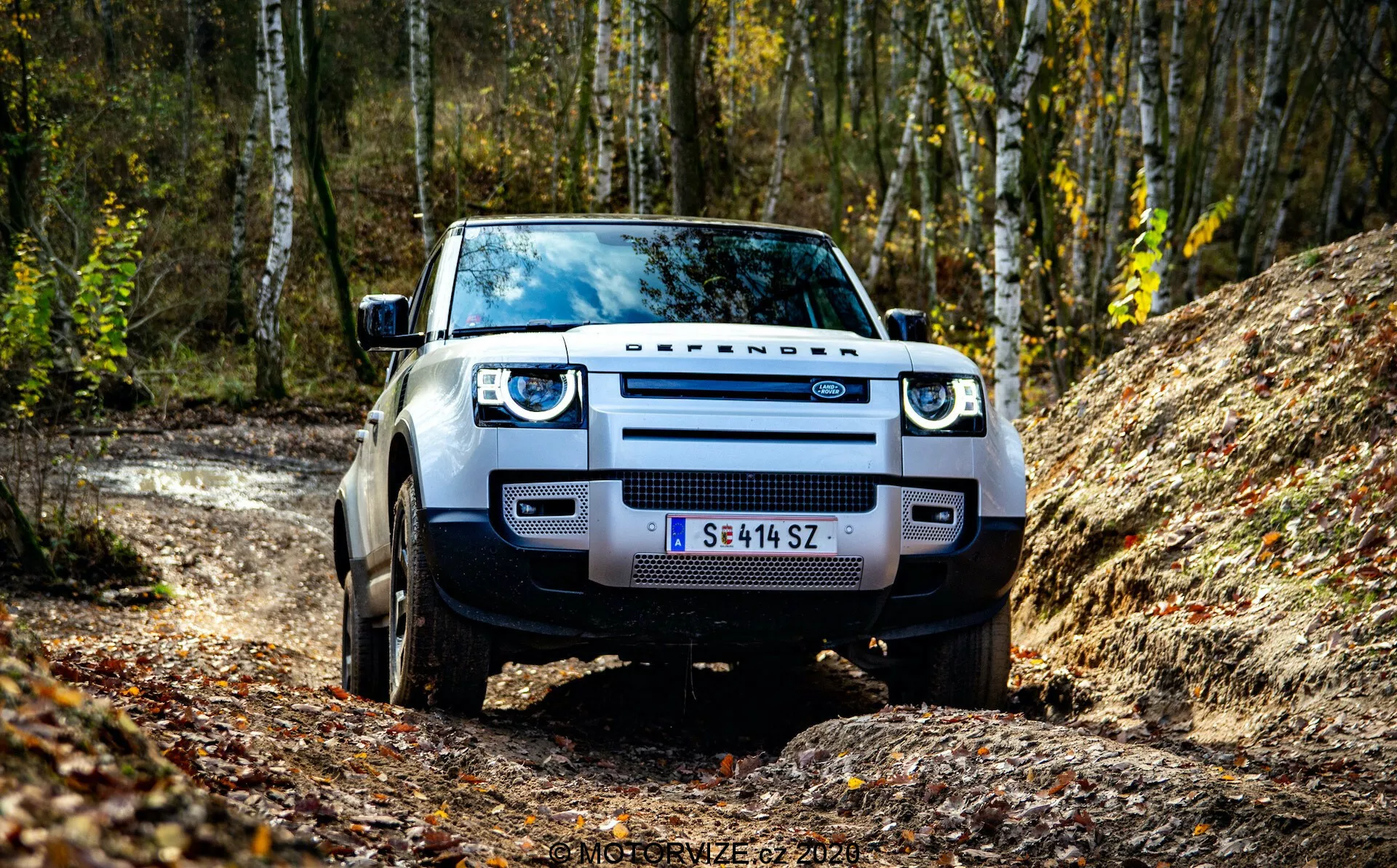 Sprednji pogled na Land Rover Defender 2020, posnet iz nizkega kota, ki poudarja njegovo rešetko, žaromete in sprednji odbijač. SUV je postavljen na neasfaltirani cesti z gozdom v ozadju, kar poudarja njegovo pripravljenost za teren. Vidni elementi vključujejo izrazit napis 'DEFENDER', LED žaromete, široke blatnike in robustne pnevmatike.