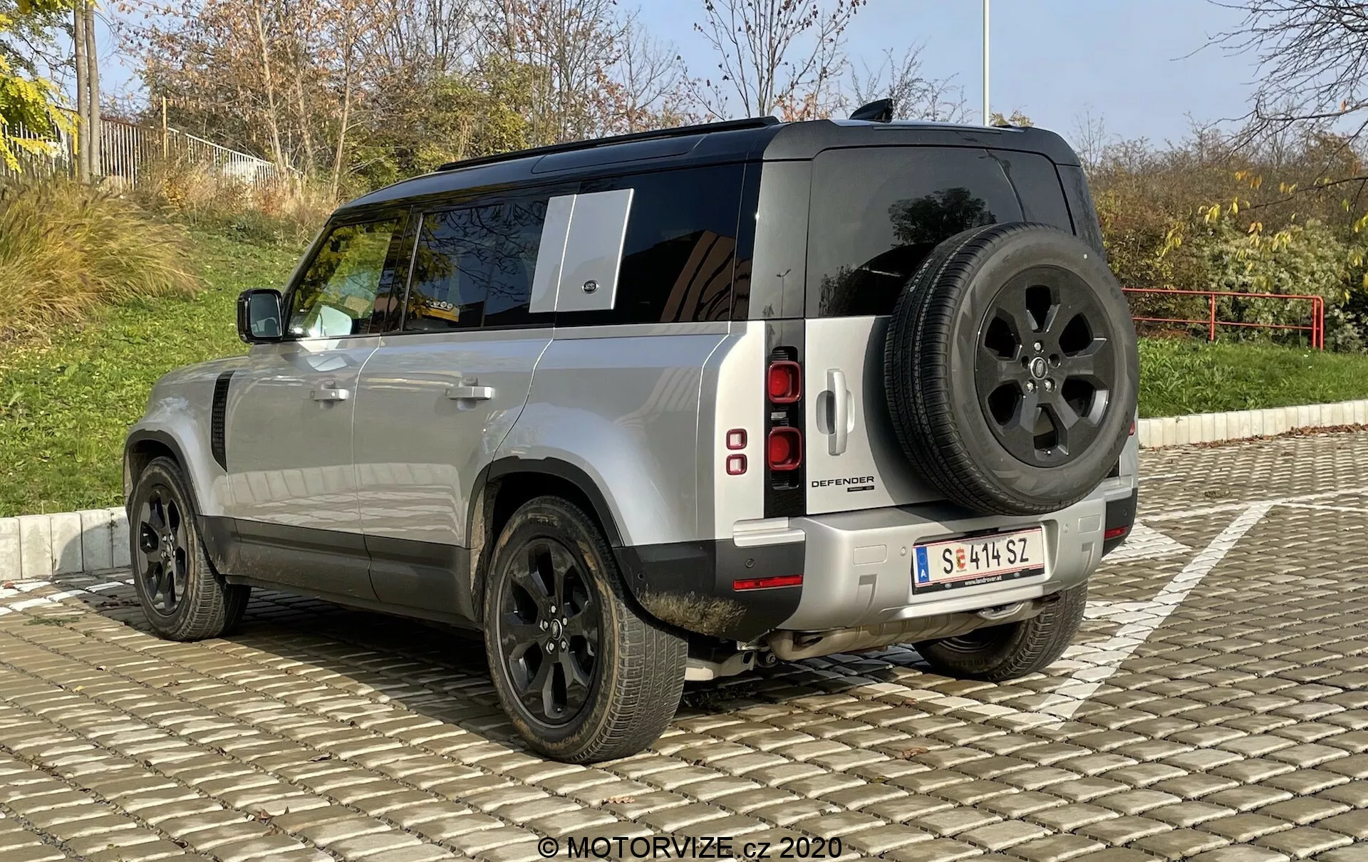 Slika prikazuje zadnji tridelni pogled na SUV Land Rover Defender iz leta 2020. Ta kot omogoča jasen pogled na škatlast, robusten dizajn vozila, kovinsko srebrno barvo in navpično segmentirane zadnje LED luči. Opazni detajli vključujejo rezervno kolo, nameščeno na zadnjih vratih, robusten zadnji odbijač s senzorji za parkiranje, velika črna aluminijasta platišča z debelimi stranskimi steni za terensko uporabo in zatemnjena zadnja stekla. Poleg tega je na strehi vidna antena, ki nakazuje na prisotnost naprednih komunikacijskih sistemov. Avto je parkiran na tlakovanem območju pod jasnim nebom.