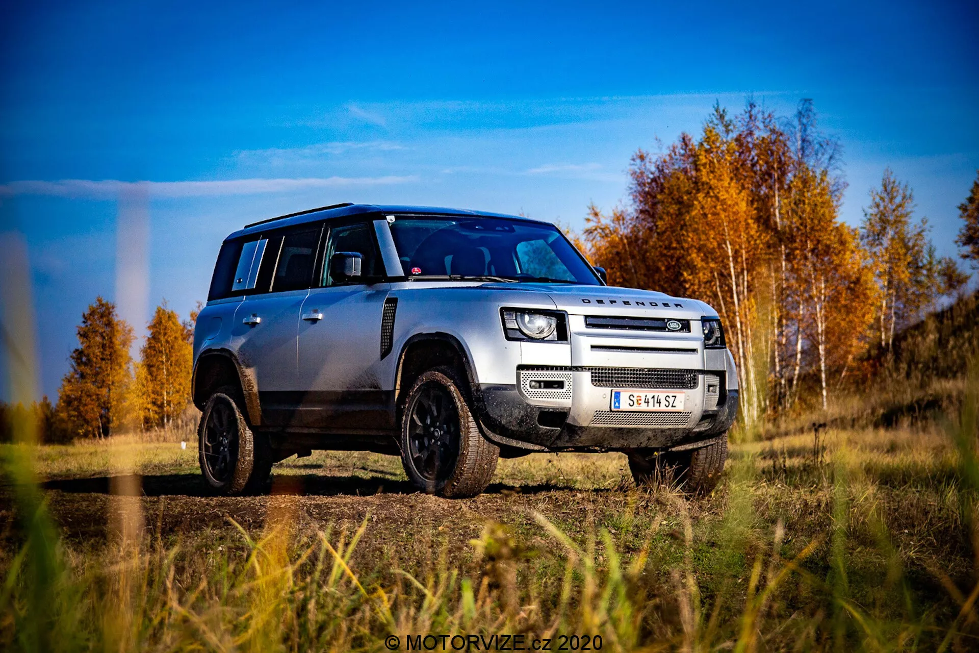 Slika srebrnega Land Rover Defender 2020 iz perspektive nizke ravni, posneta v nizkem, poševnem prednjem poglednem kotu v treh četrtinah. Vozilo jasno kaže svojo mrežo, pokrov motorja, sprednji odbijač in dizajn koles, rahlo usmerjeno proti voznikovi strani. Defender je na zunanjem terenu s podlago jesenskih dreves z rdečimi in oranžnimi listi pod modrim, delno oblačnim nebom. Poudarjene oblikovne lastnosti vključujejo izrazito škatlasto obliko, moderne LED žaromete, meglenke, dvobarvno mrežo z blagovno znamko LAND ROVER, sprednjo zaščitno ploščo, črno aluminijasta platišča z terenskimi pnevmatikami in kontrastno črno streho z ujemajočimi se črnimi pokrovi ogledal. Fotografija je zelo ostra in dinamična, poudarjajoč robustnost in moderno estetiko vozila.