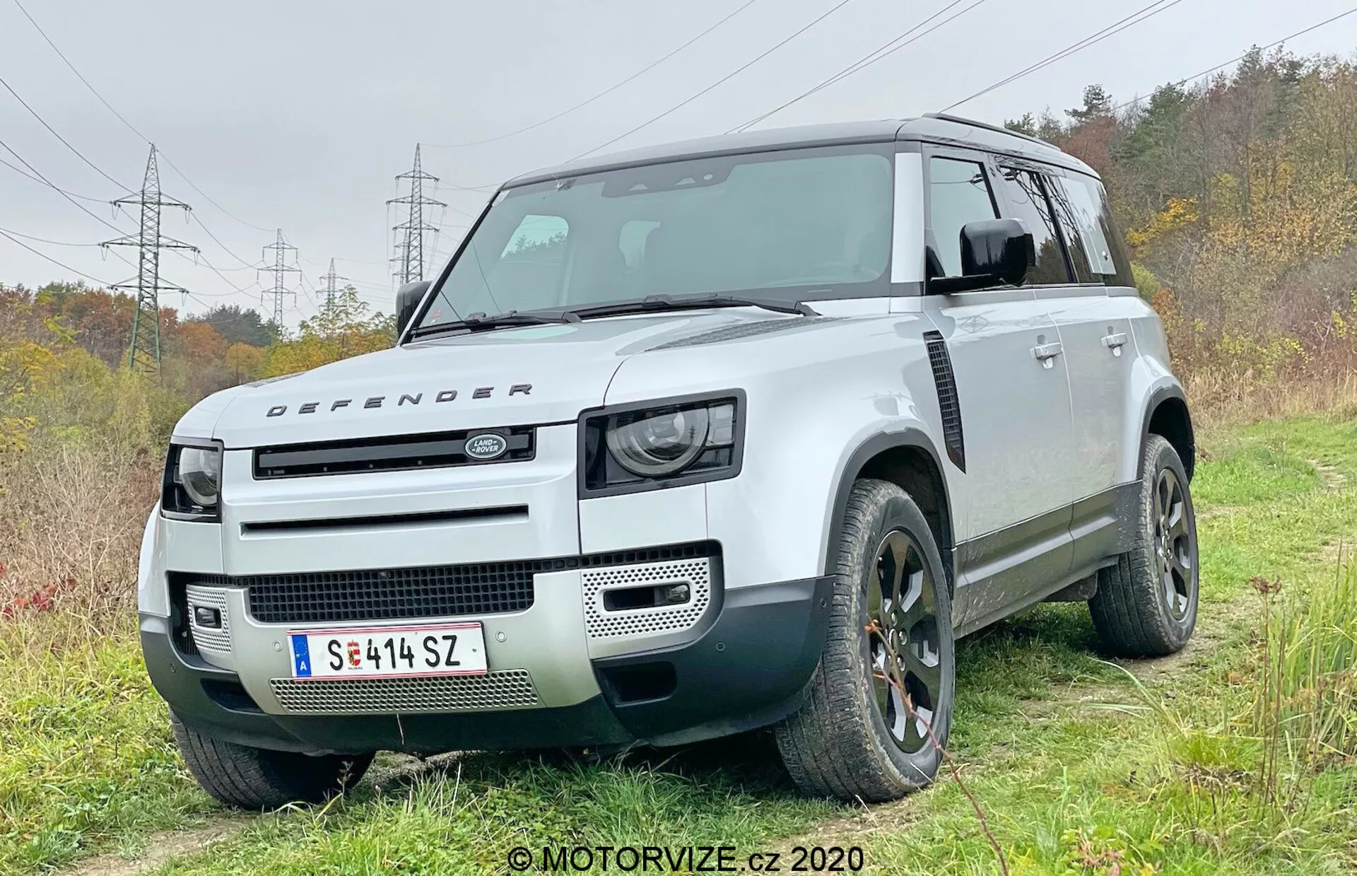 Ta fotografija zajema moderno SUV vozilo Land Rover Defender 2020 iz sprednjega četrtinskega kota, ki prikazuje voznikovo stran. Vozilo je rahlo na makadamski cesti na travnatem območju, kar poudarja njegove terenske zmogljivosti. Značilna škatlasta zasnova z visoko oddaljenostjo od tal, dvobarvna barvna shema z belo karoserijo in kontrastno črno streho. Sprednji del prikazuje retro navdihnjene LED žaromete, pokrov motorja z značko Defender in minimalistično sprednjo rešetko, kar poudarja svojo robustno terensko slovest. Vidne tehnične funkcije vključujejo sprednje parkirne senzorje, vgrajene v odbijač, kar nakazuje napredne sisteme za pomoč vozniku. Močni okvirji koles sprejemajo velike terenske pnevmatike, in oblikovalski elementi, kot so aluminijasta platišča in edinstvena zasnova rešetke, so poudarjeni.