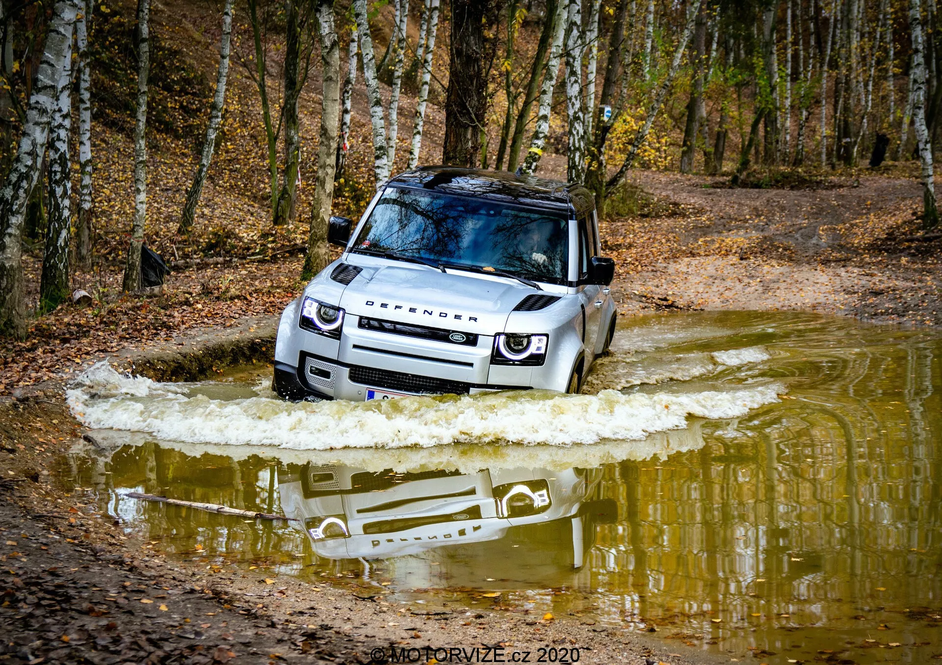 Posnetek iz nizkega kota prikazuje Land Rover Defender 2020, ki prečka vodno telo, pri čemer izpostavlja sprednjo desno stran vozila z vidno mrežo, LED žarometi, pokrovom, vetrobranskim steklom, streho, sprednjim kolesom in voznikovimi vrati. Dinamični prizor zajame premikanje vode, ki poudarja terenske sposobnosti, visoko zmožnost prečkanja vode, zaščitno ploščo in luksuzno panoramsko streho. Znamka Land Rover je vidna na pokrovu motorja, kar kaže na robustno in vsestransko zasnovo.