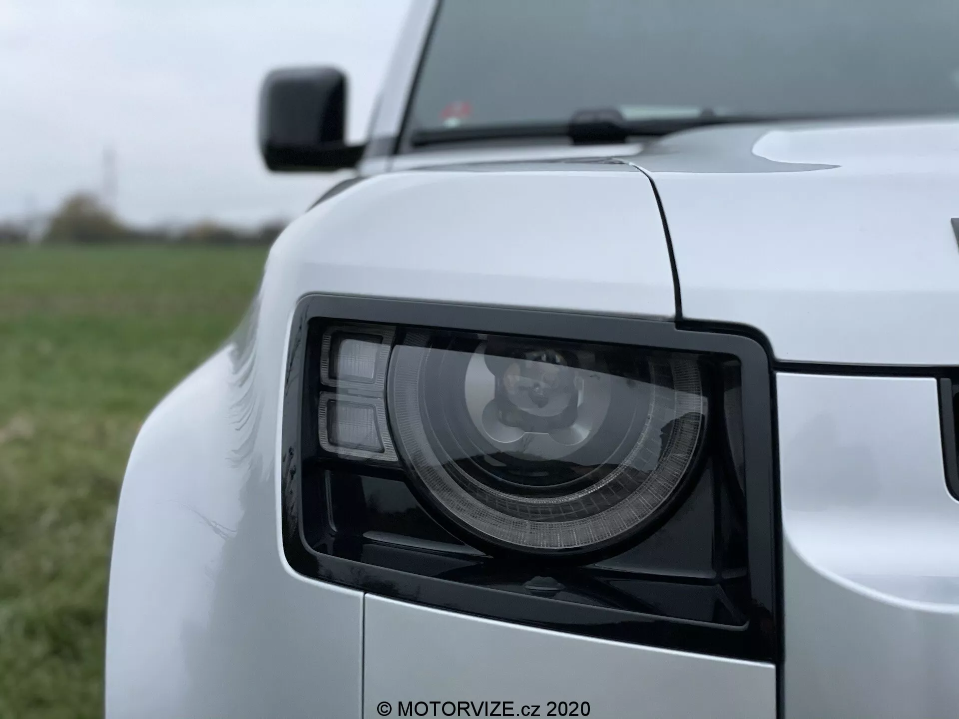 Pogled od blizu levo-sprednje luči Land Rover Defender 2020 z modernim kotnim dizajnom, posnet pod rahlim kotom in nizko-srednjo višinsko perspektivo. Slika prikazuje podrobne LED/HID svetlobne module, sijajni beli lak ter del neizostrene sprednje rešetke, roba pokrova motorja, sprednje strani odbijača in vzvratnega ogledala.