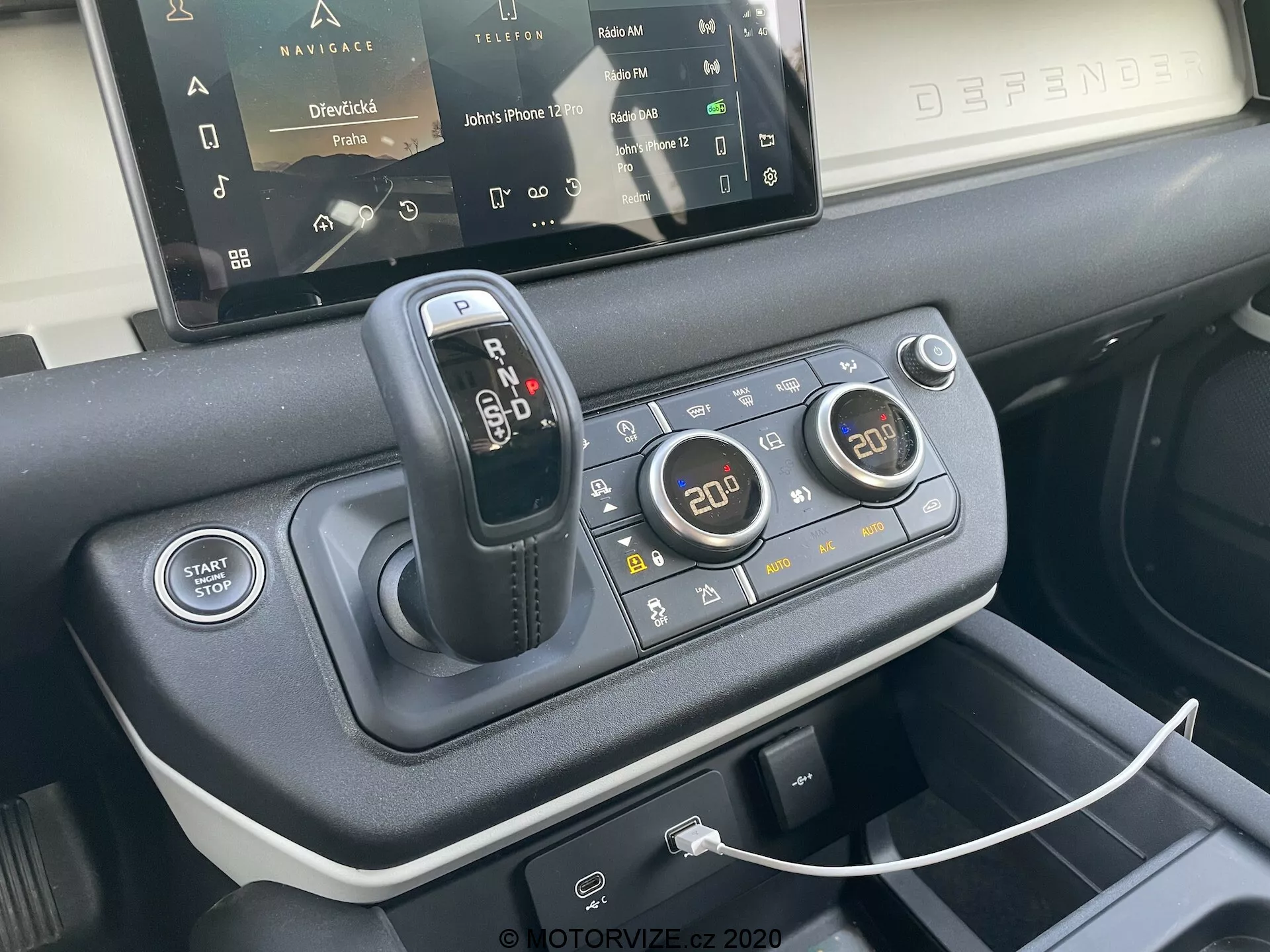 Pogled od blizu na sredinsko konzolo modela Land Rover Defender 2020, ki poudarja prestavno ročico, terenski odzivni sistem za različne vozne pogoje, gumb za zagon/ustavitev motorja, dvopodročno klimatsko napravo, USB priključek za povezljivost naprav, delno viden zaslon na dotik infotainment sistema in vrhunske materiale, uporabljene pri oblikovanju notranjosti, gledano s perspektive sprednjega sedeža pod kotom 45 stopinj.