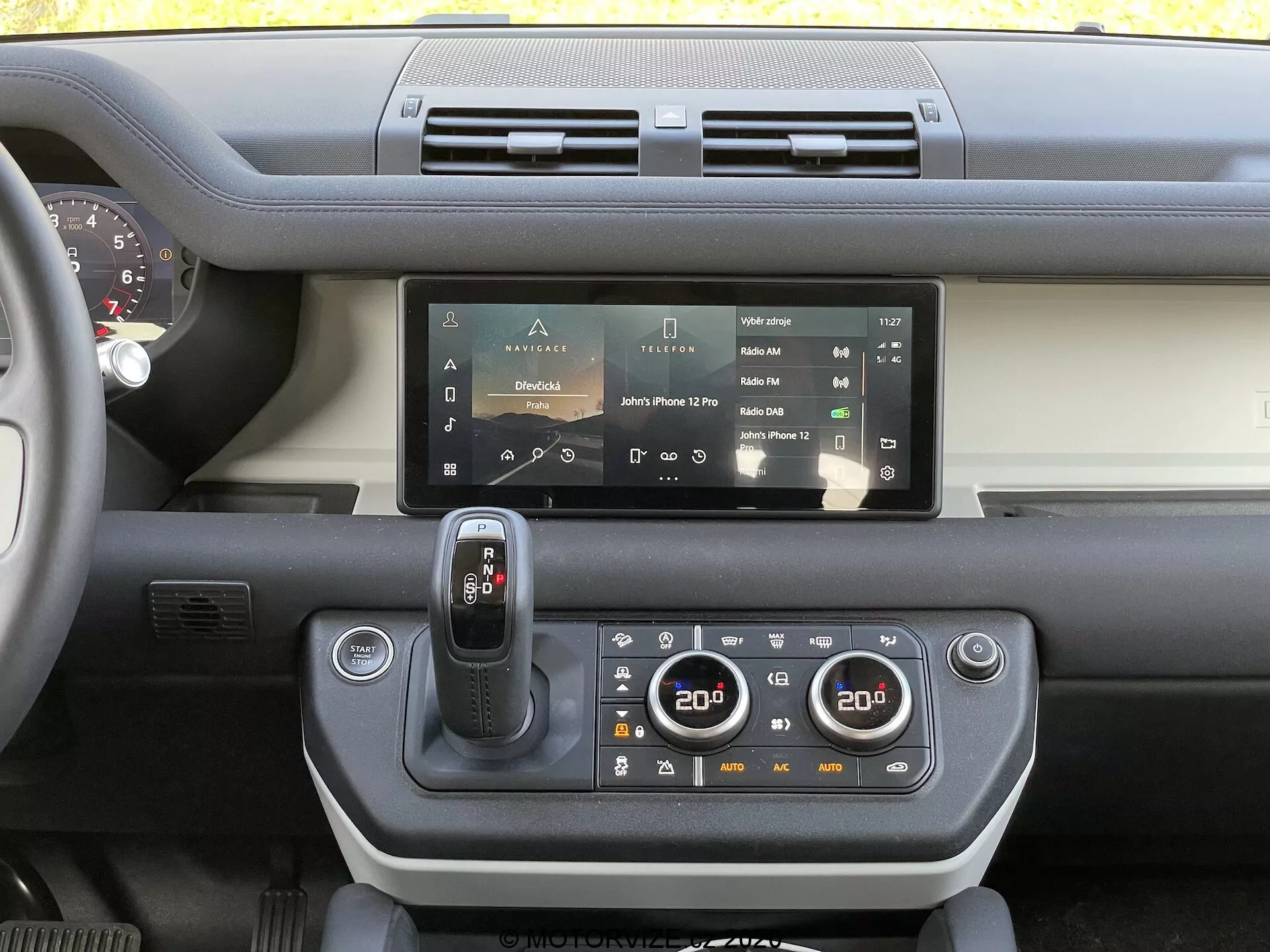 Notranji pogled na armaturno ploščo Land Rover Defender 2020, ki prikazuje zaslon na dotik infotainment sistema, dvojne zračne odprtine, sodoben gumb za izbiro prestav, dvopodročno klimatsko nadzorno ploščo, pogled na volan in kombinacijo trdih plastičnih ter mehkih materialov z barvnim kontrastom na armaturni plošči.