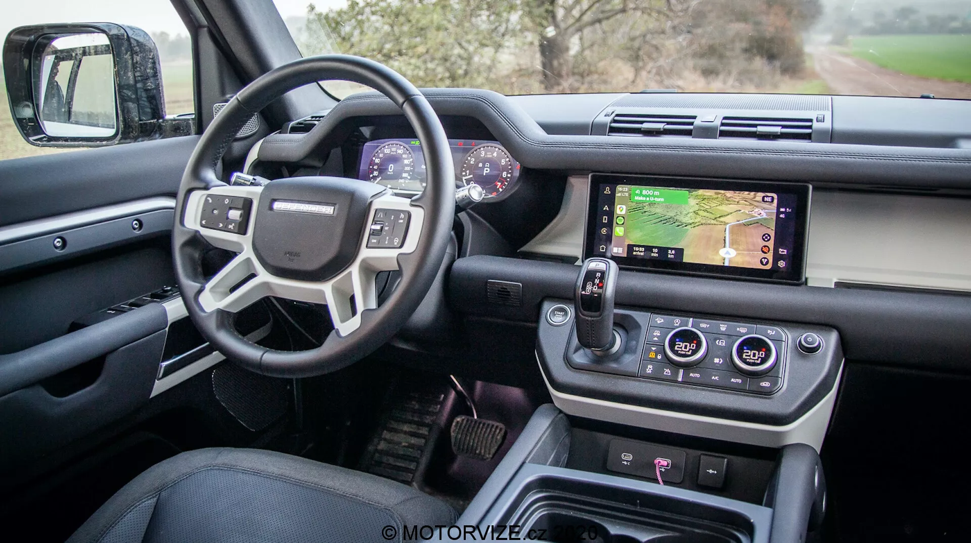 Notranji pogled na moderni Land Rover Defender 2020 z voznikove strani vrat, ki prikazuje sofisticiran dizajn in napredne funkcije, vključno z odprtimi voznikovimi vrati z nadzorom oken in ogledal, večfunkcijskim volanom z nadzornimi gumbi, digitalnim instrumentnim zaslonom, osrednjim infotainment sistemom z zaslonom na dotik, ki prikazuje zemljevid, gumbi za nadzor klime, sodobnim rotacijskim izbirnikom prestav in gumbi za nadzor terena, vse v luksuzni visoko kakovostni izvedbi.