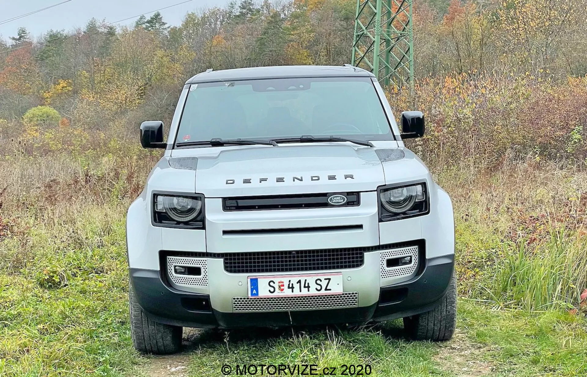 Frontalni pogled na bel Land Rover Defender 2020 4x4, z izrazito pravokotno mrežo, obdano s krožnimi LED žarometi s vgrajenimi kvadratnimi dnevnimi lučmi, robustnim sprednjim odbijačem s pravokotnimi režami, ravnim pokrovom z napisom 'LAND ROVER' in oznako 'DEFENDER' na sprednjem robu, ravnim vetrobranom, osrednje nameščeno evropsko registrsko tablico in parkirnimi senzorji, zajeto v naravnem okolju.