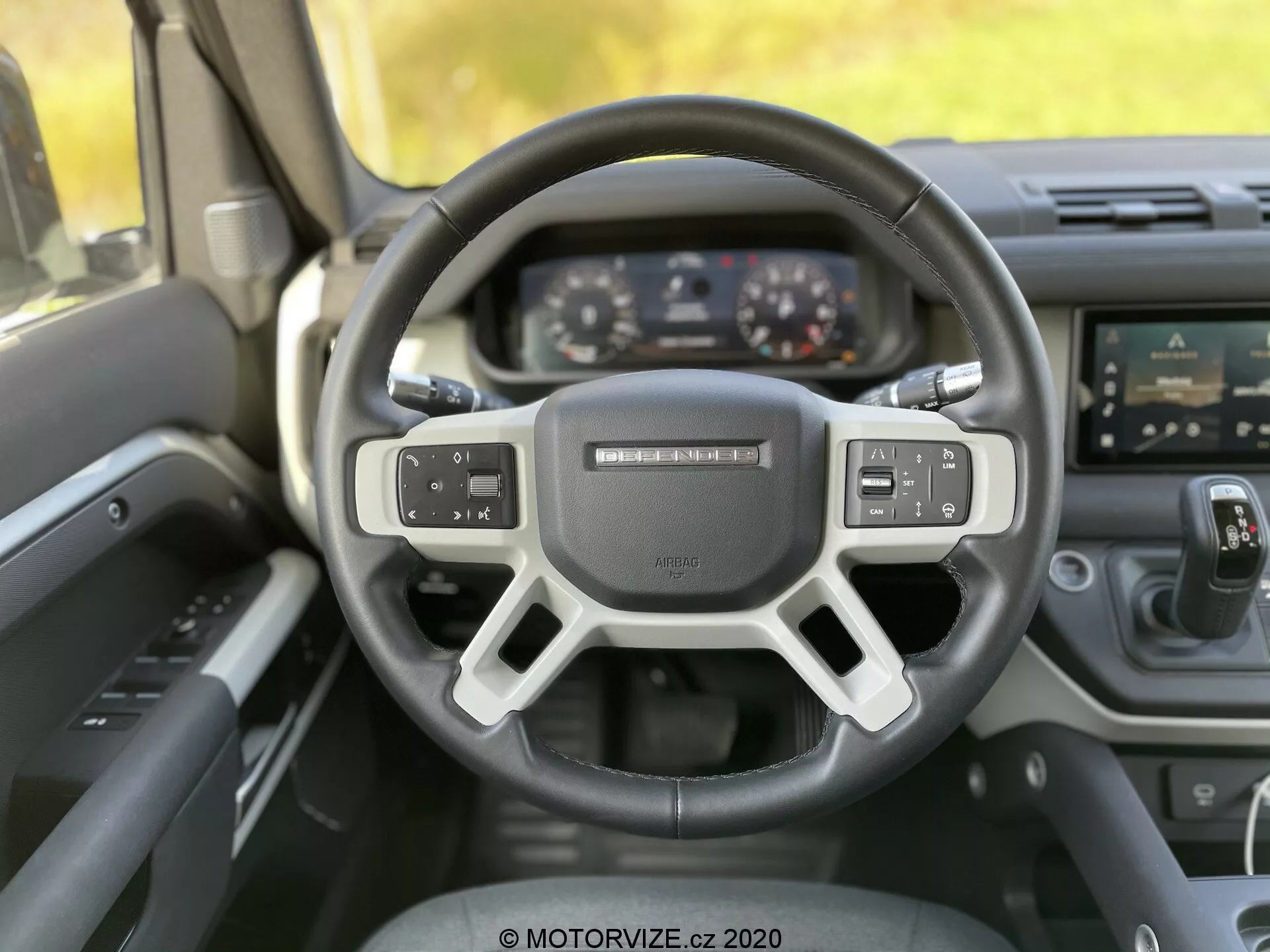 Pogled na notranjost Land-Rover Defender 2020 iz perspektive voznika, s sodobnim trikrako volanom z vgrajenimi kontrolami za zvočne nastavitve, glasovne ukaze in nastavitve tempomata. Za volanom je razvidna digitalna instrumentna plošča z zasloni za merilnik hitrosti, tahometer in večfunkcijski zaslon. Slika prav tako prikazuje sredinsko konzolo z zaslonom na dotik za infotainment sistem in spodnjimi gumbi za nadzor temperature. Kot fotografije je rahlo nagnjen navzgor, poudarjajoč ergonomski dizajn, napredno tehnologijo in kakovostne materiale stacionarnega vozila.