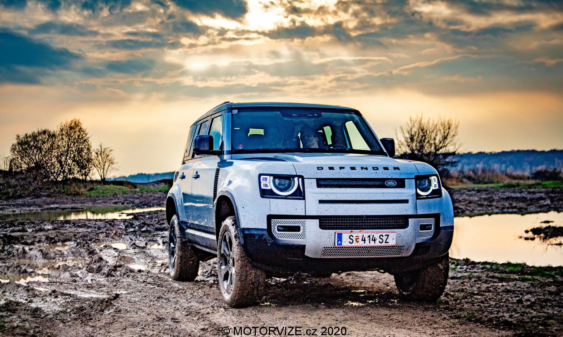 Fotografija SUV Land Rover Defender posneta iz sprednjega, rahlo izven osrednjega kota, poudarjajoč sprednjo fasado vozila in njegovo terensko zmogljivost. Vozilo je svetle barve, z umazanijo na pnevmatikah in spodnjih delih karoserije, kar kaže, da je bilo voženo po terenu. Avto je opremljen s šnorklom za prečkanje globoke vode in LED žarometi. Podeželsko okolje vključuje blatnat teren in stoječo vodo, z dramatičnim zgodnje večernim nebom v ozadju.