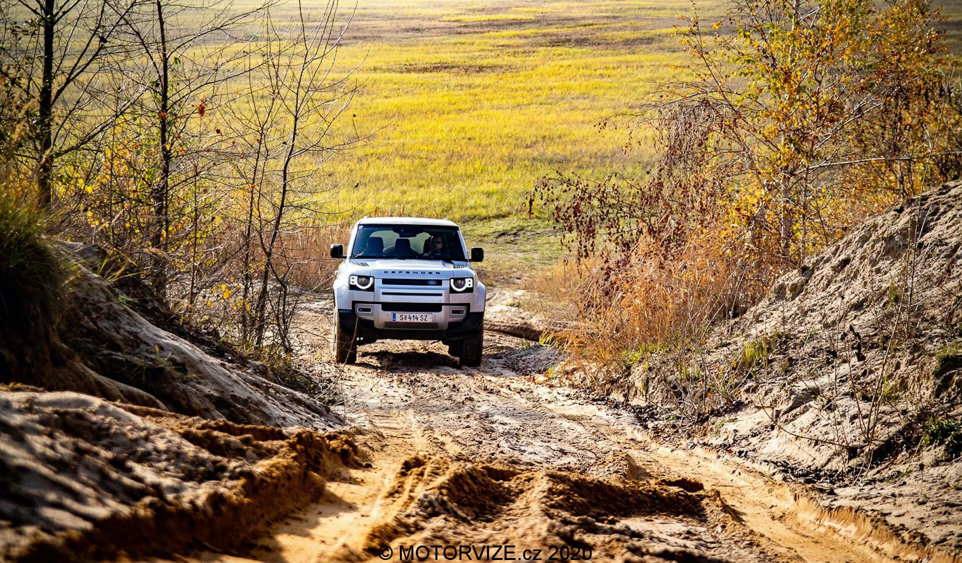 SUV Land Rover Defender 2020 je videno od spredaj vožnjo po terenu, nameščen na sredini proti ozadju neravnega terena in naravnega okolja. Vidni sprednji del avtomobila prikazuje napredno artikulacijo vzmetenja, LED žaromete in značilno rešetko z vodoravnimi letvicami. Zajemanje SUV v podeželskem okolju z jesenskimi odtenki in naravno svetlobo, perspektiva poudarja robustno in pustolovsko prisotnost vozila.