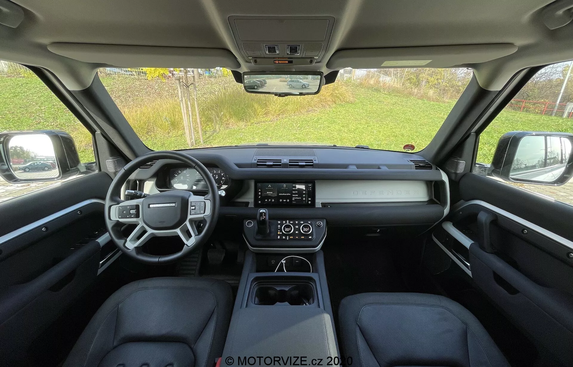 Pogled na notranjost Land Rover Defender 2020 s zadnjega sedeža, ki prikazuje eleganten, sodoben kabinet z večfunkcijskim volanom, vodoravno armaturno ploščo s centralnim digitalnim zaslonom, vidnim zaslonom na dotik infotainment sistemom, dvopodročno klimatsko napravo in čisto sredinsko konzolo z elektronskim prestavljalnikom in držali za kozarce.